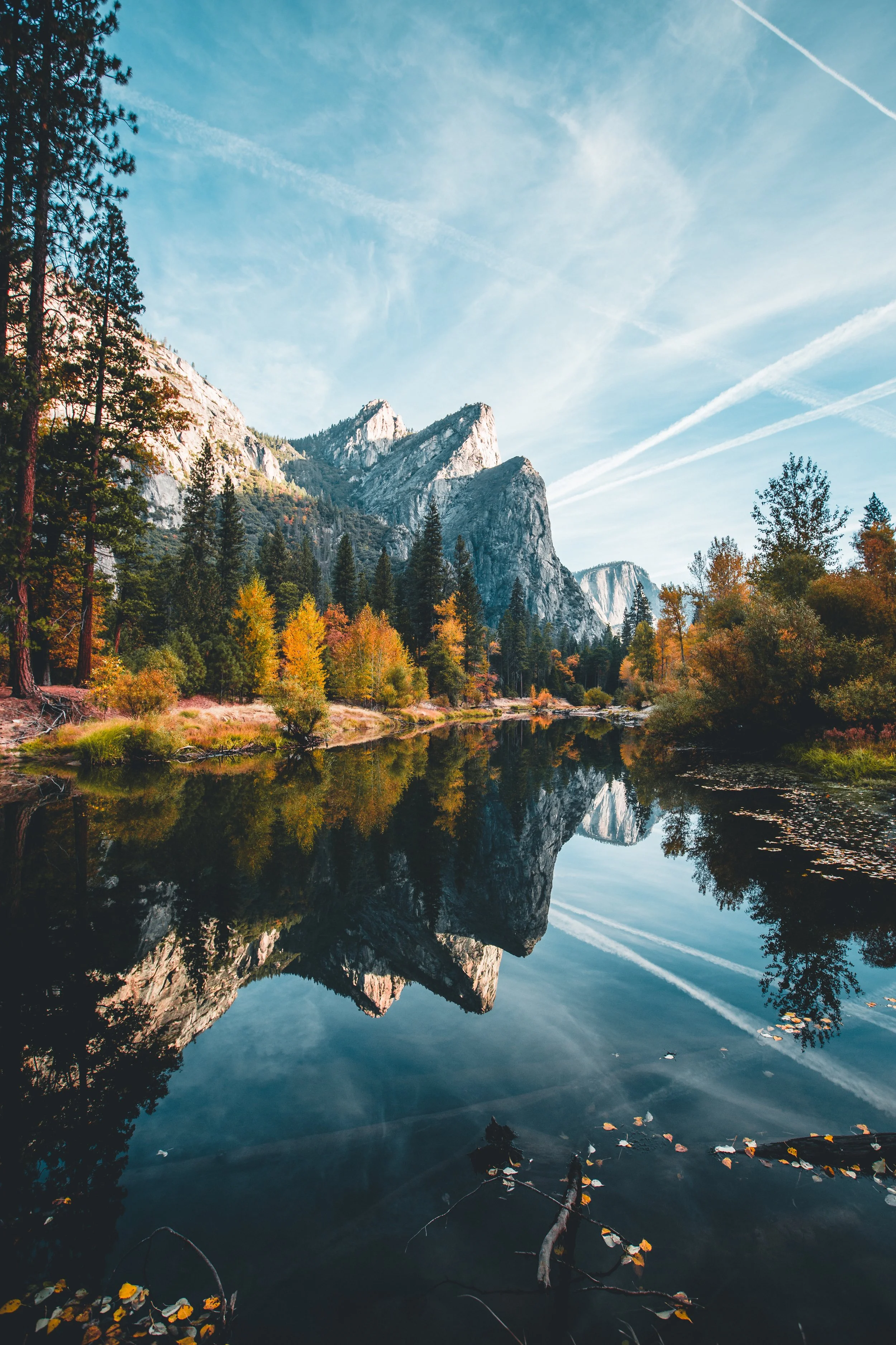 A peaceful mountain scene with tall rocky peaks, a calm reflective river, colorful autumn trees, and a bright sky with white clouds and contrails.