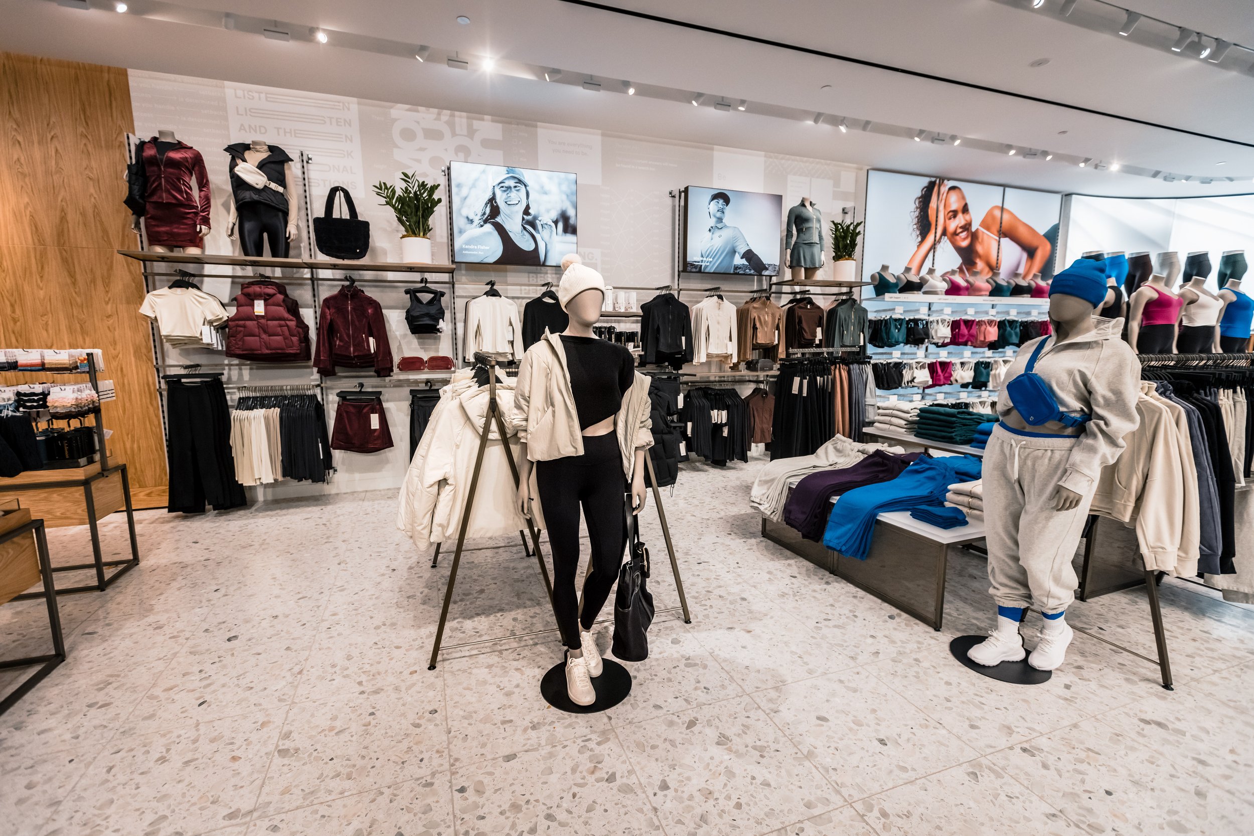 Inside a retail clothing store with mannequins dressed in activewear, shelves with folded clothes and jackets, and electronic screens displaying fitness models.