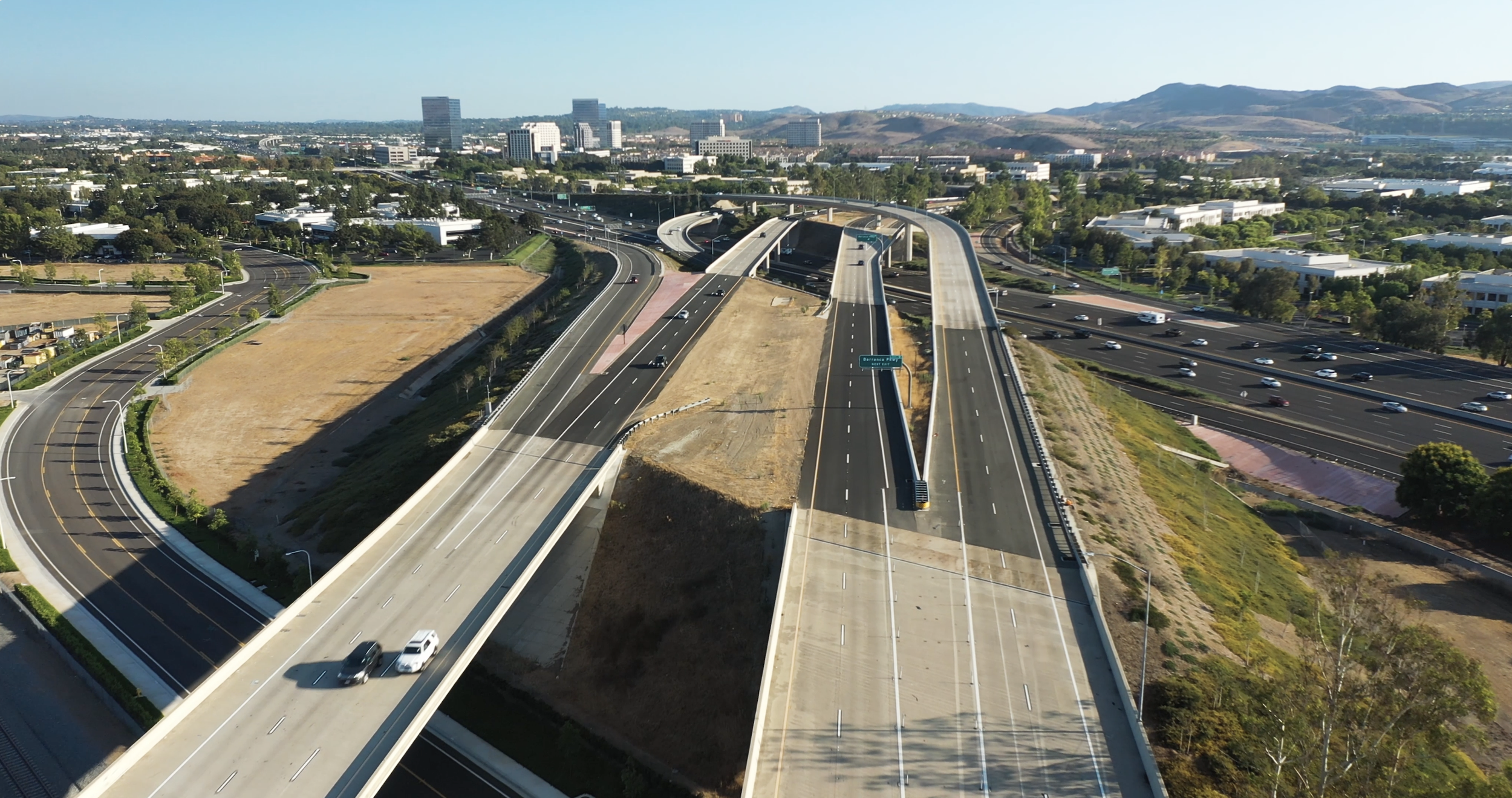Elevated view of multiple highways and overpasses with few cars in a cityscape, surrounded by greenery and distant hills.
