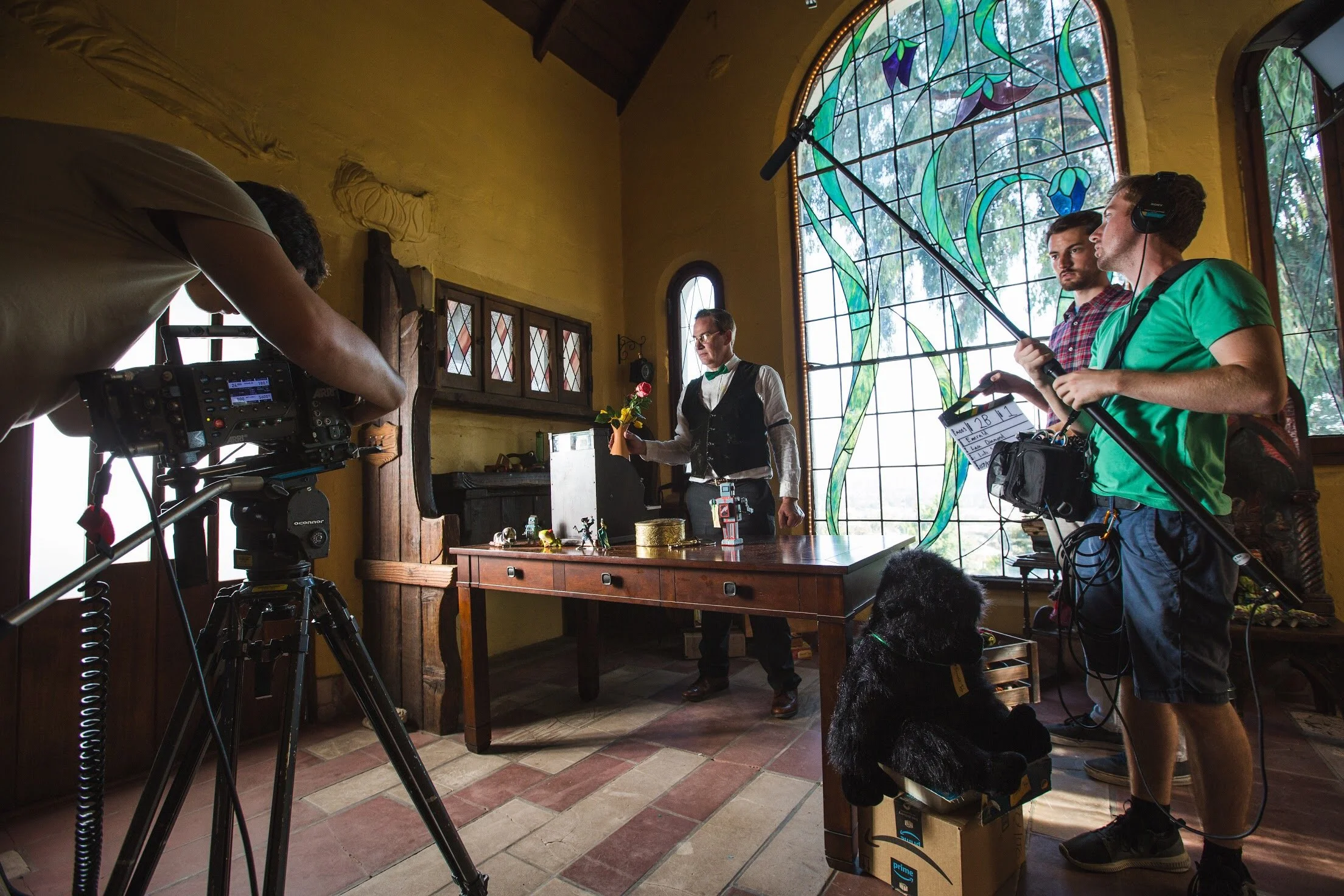 Film crew filming a scene inside a room with yellow walls and a large stained glass window, person at the table holding a red rose, camera operator adjusting the camera, and the director with headphones, a boom mic, and a clapperboard.