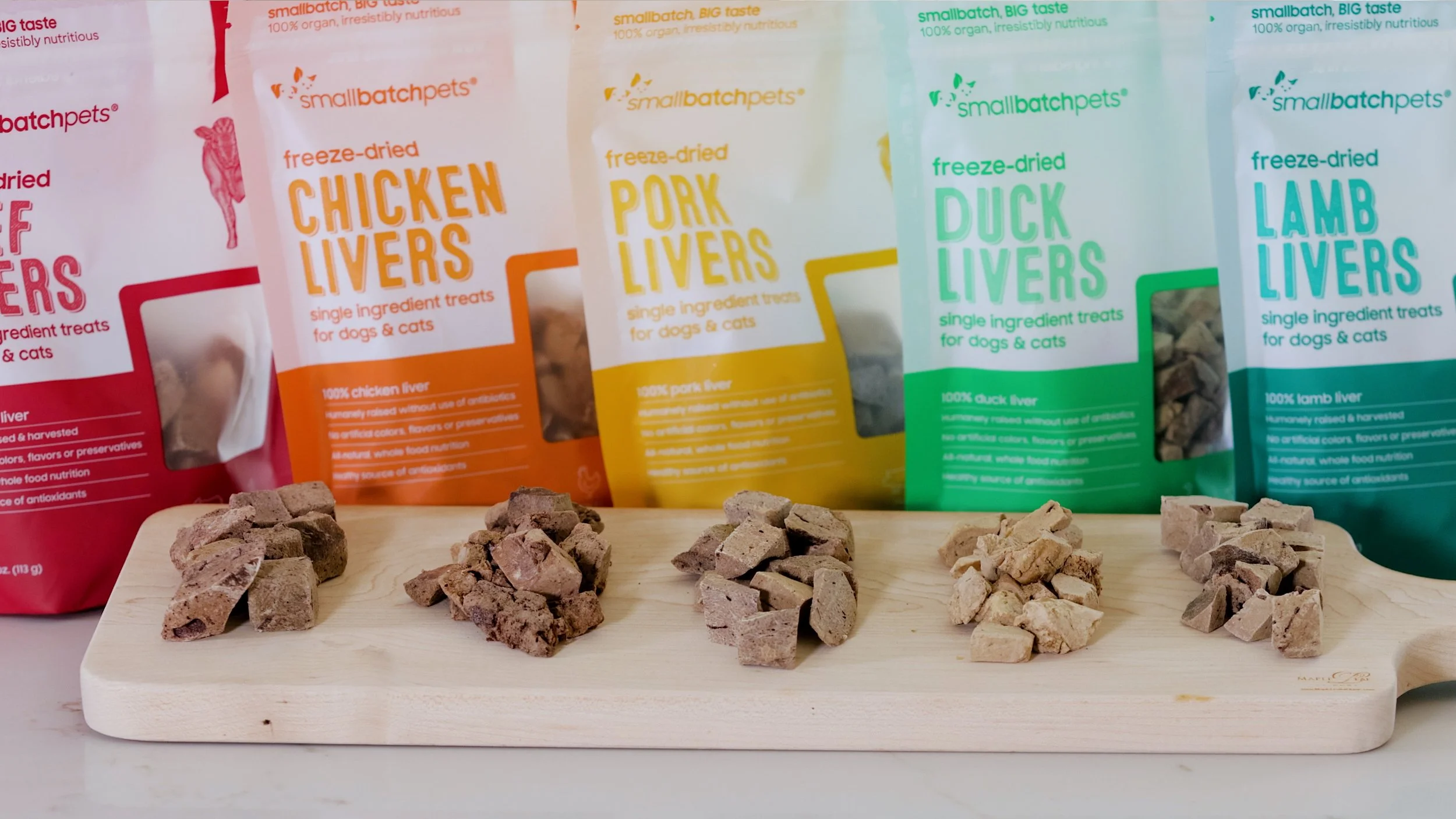 Four samples of freeze-dried dog treats, including chicken liver, pork liver, duck liver, and lamb liver, arranged on a light wooden board in front of colorful packages of the same treats.