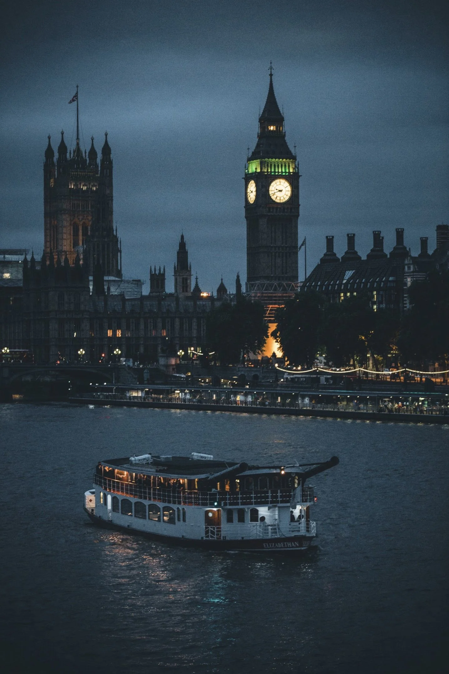 Nighttime view of London skyline with Big Ben clock tower, Houses of Parliament, and a boat on the River Thames.
