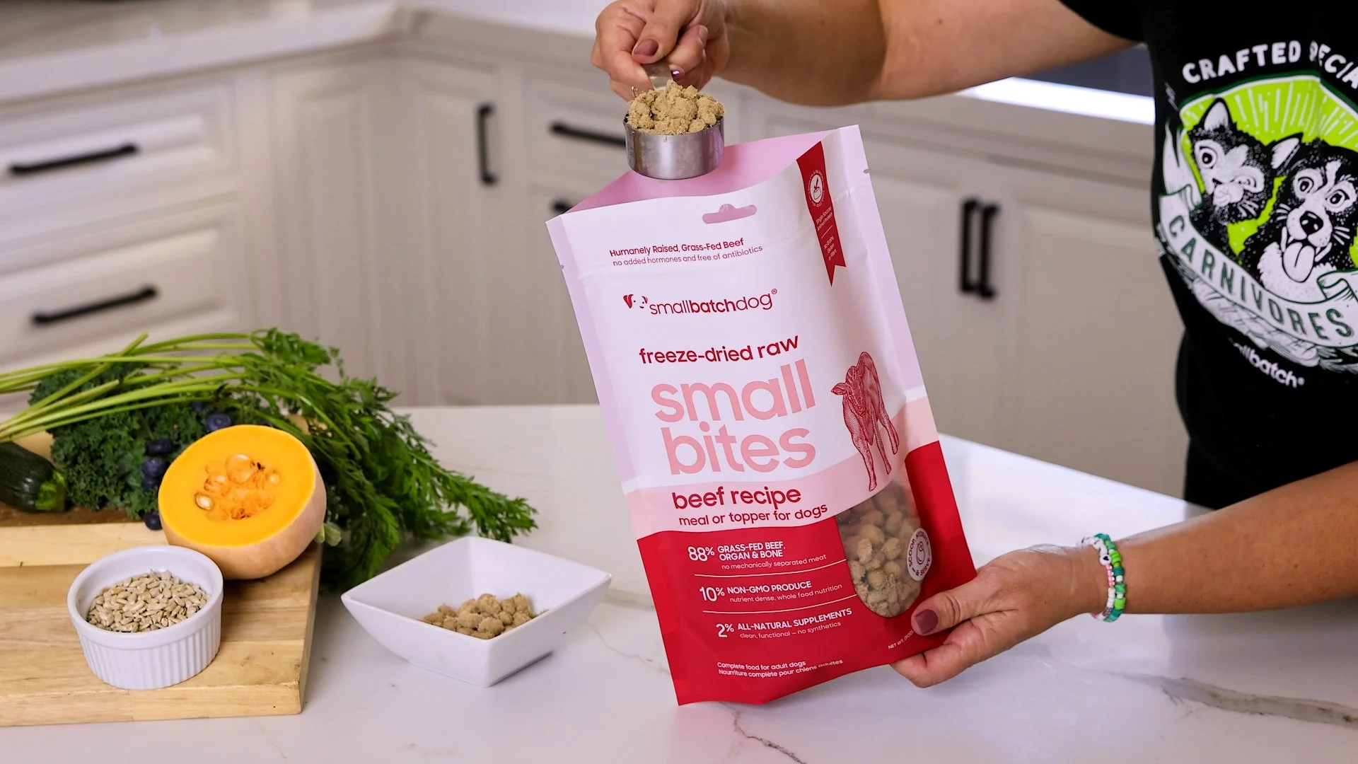 Person holding a package of small bites freeze-dried raw beef treats for dogs in a kitchen. There are fresh vegetables and seeds on the counter.