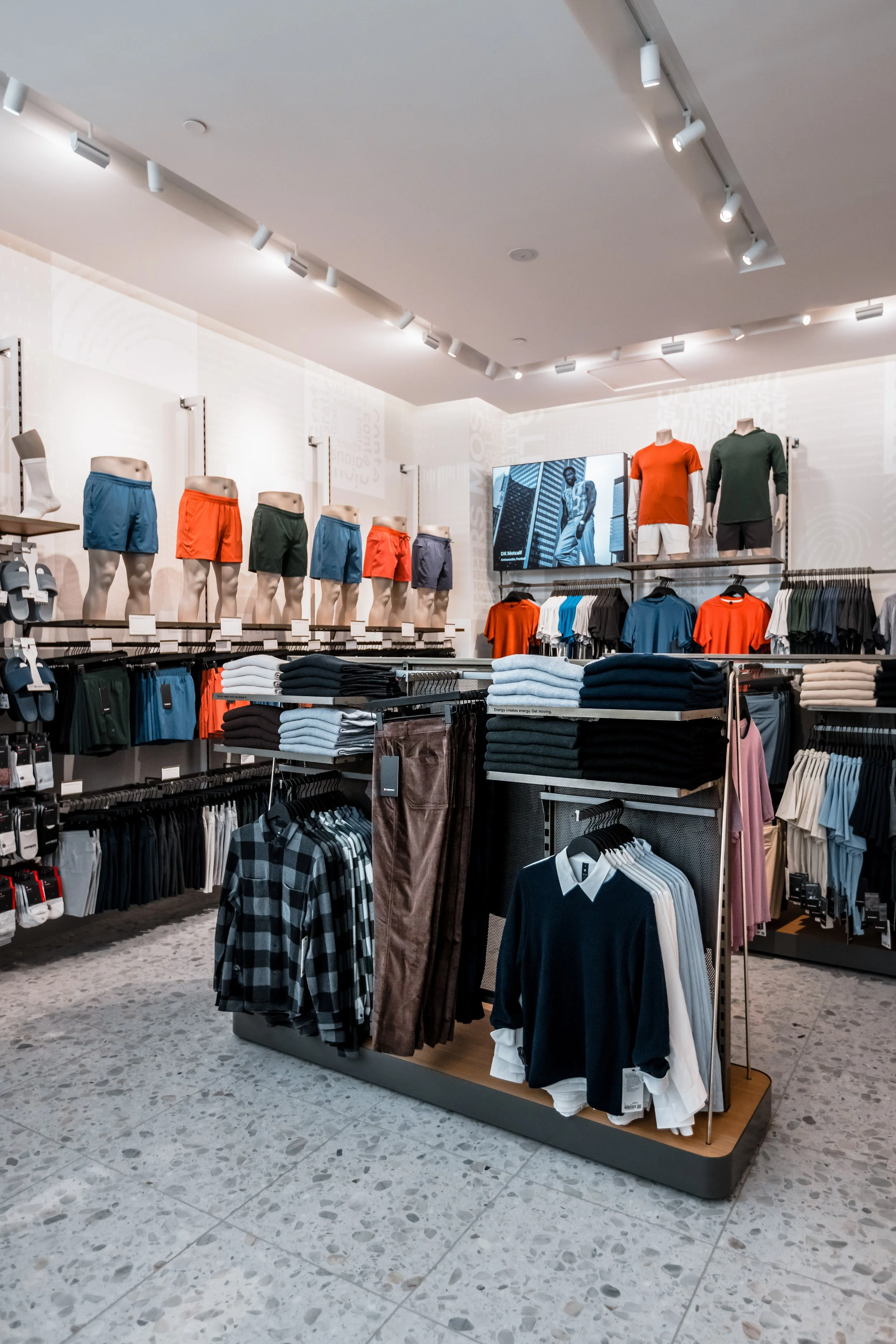 Clothing store display with mannequins in shorts and t-shirts, shelves of folded clothes, and racks of shirts and pants.