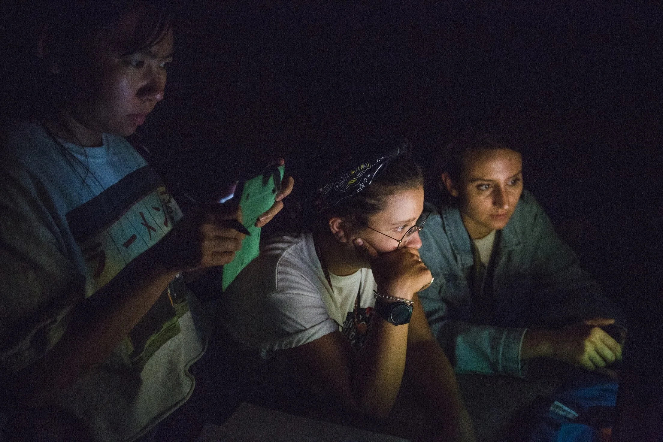 Three people sitting in darkness, illuminated by a device held by the person on the left, watching something intently.