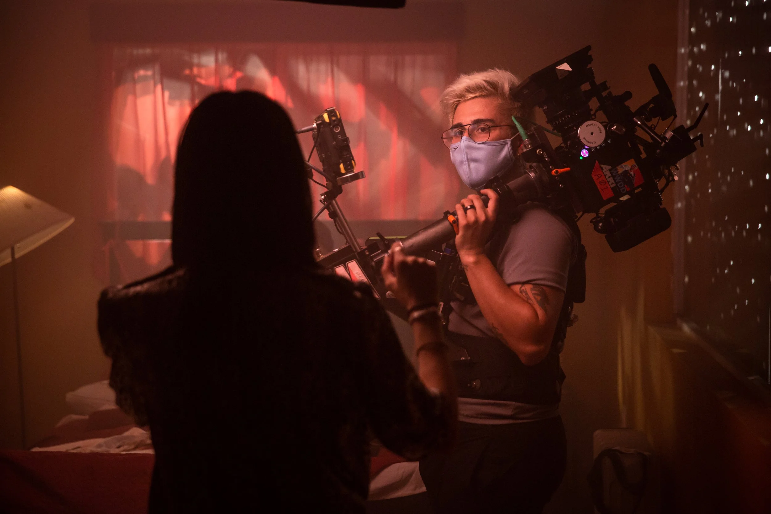 A person in a face mask holds a large camera rig, filming a woman in a dark room with red lighting.