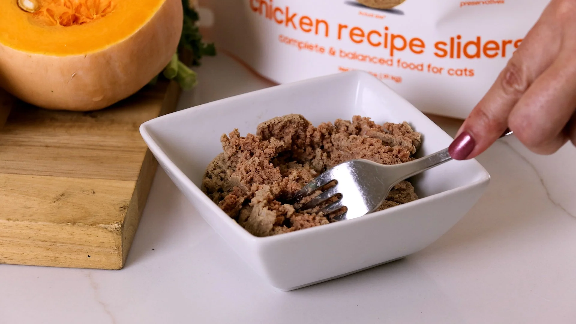 A white square bowl of canned dog food on a white surface, with a person holding a fork over it. In the background, there is a halved pumpkin and a bag of chicken recipe sliders for cats on a wooden surface.
