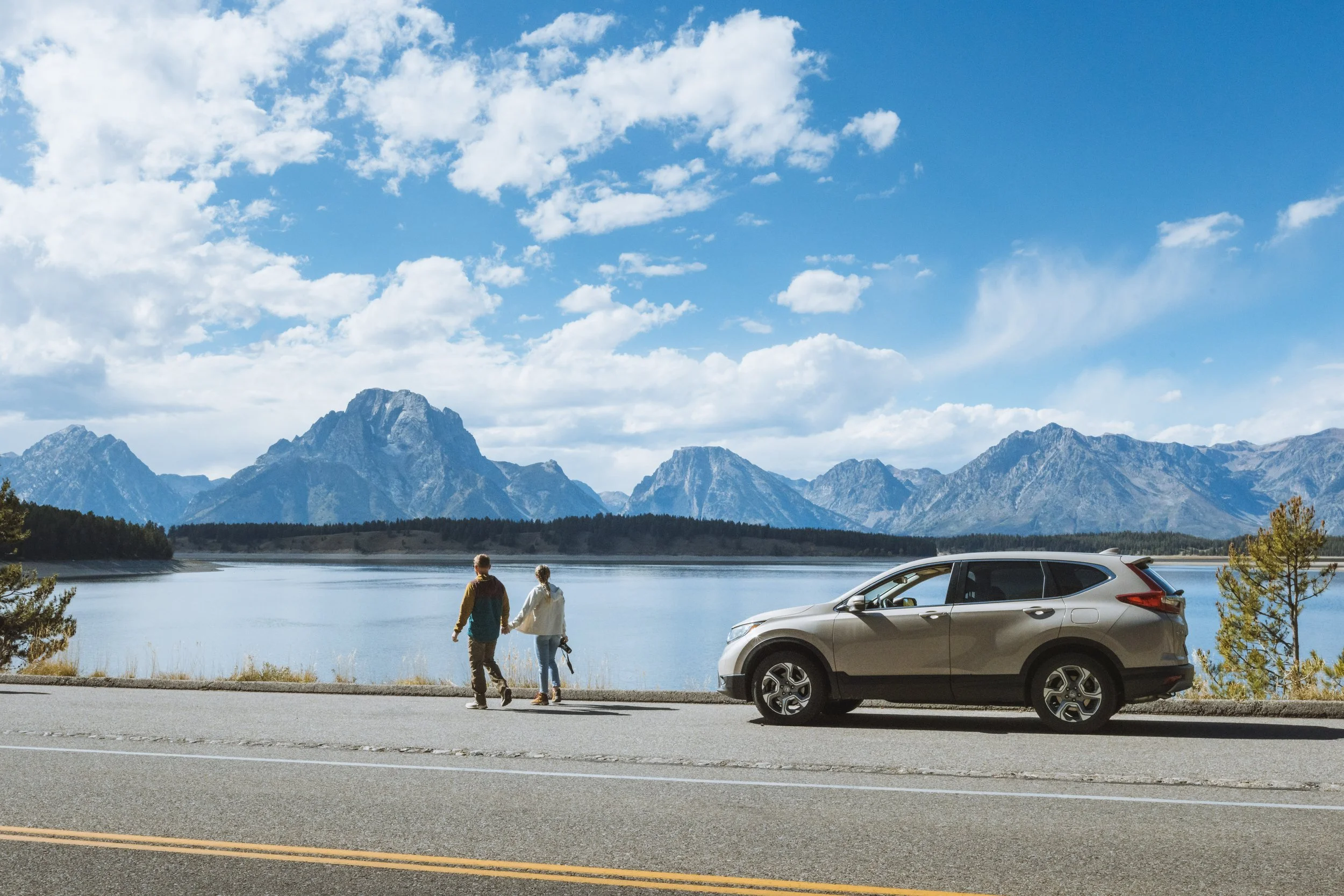 A couple walking by a lake with mountains in the background under a partly cloudy sky, parked car on the side of the road.
