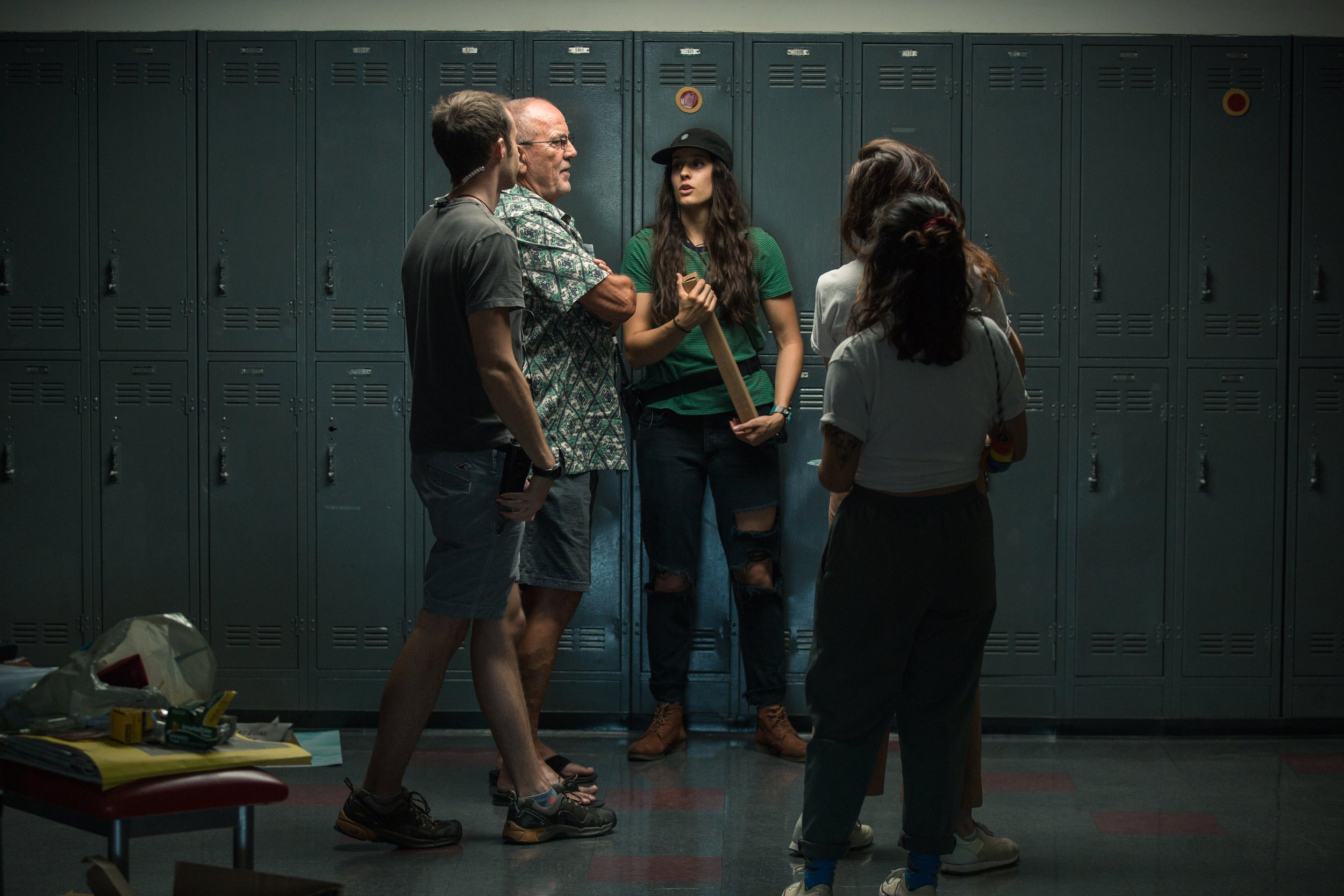 A group of five teenagers standing in a school hallway with blue lockers, having a conversation. One girl with long dark hair is holding a wooden bat, and it is evening or a dark setting.
