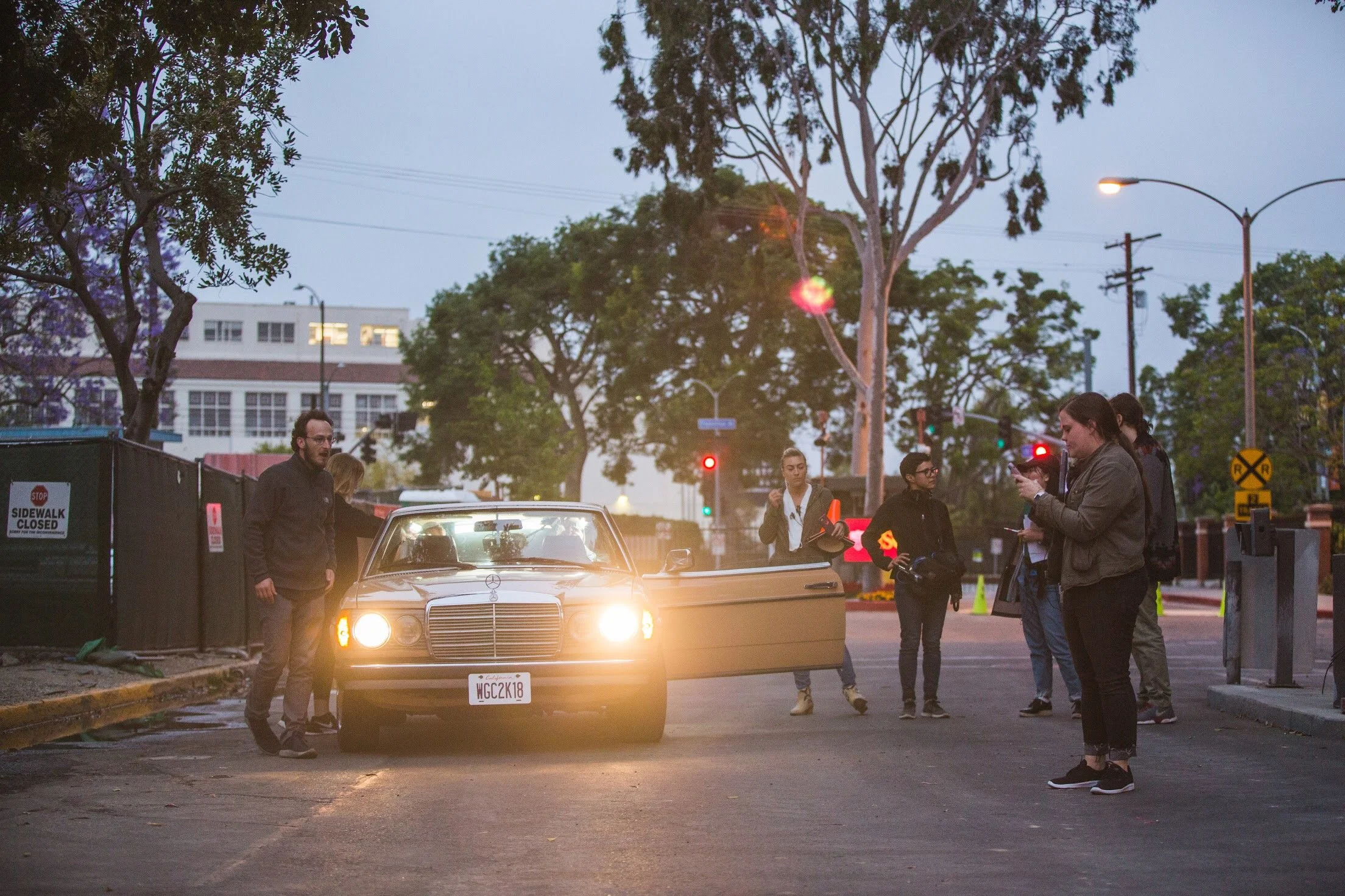 Group of people gathered around a vintage Mercedes-Benz car with its driver's side door open at dusk on a city street, some using cell phones, with trees, streetlights, and traffic signals in the background.