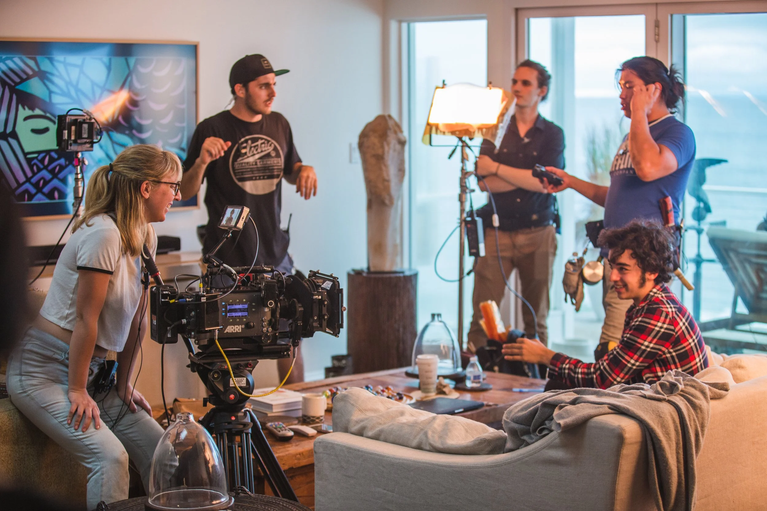 A film crew working inside a living room, capturing a scene with a young man sitting on a couch holding a snack, surrounded by production equipment and crew members.