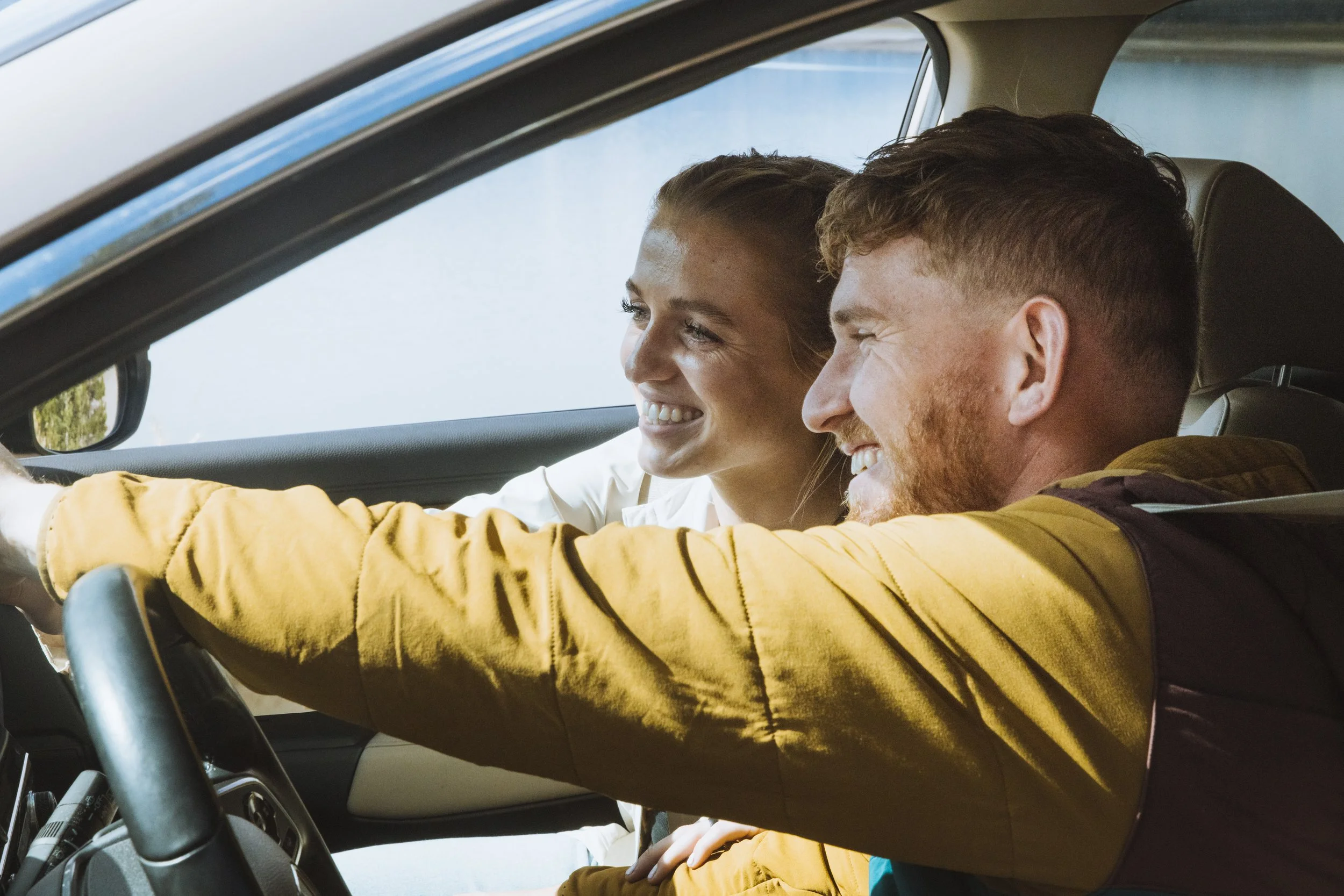 A smiling couple inside a car, with the man driving and the woman sitting next to him.