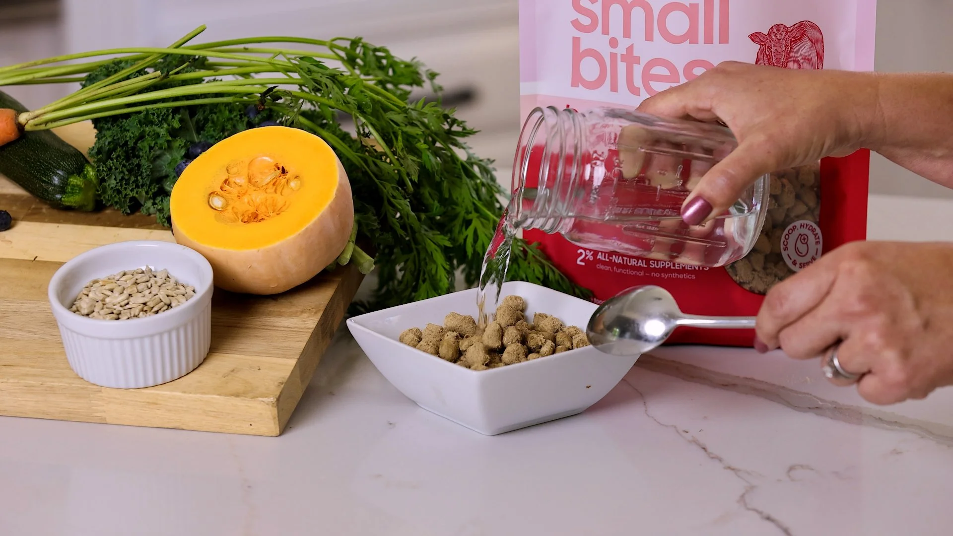 Person pouring water into a bowl of croutons on a kitchen countertop with various vegetables and a pumpkin nearby.
