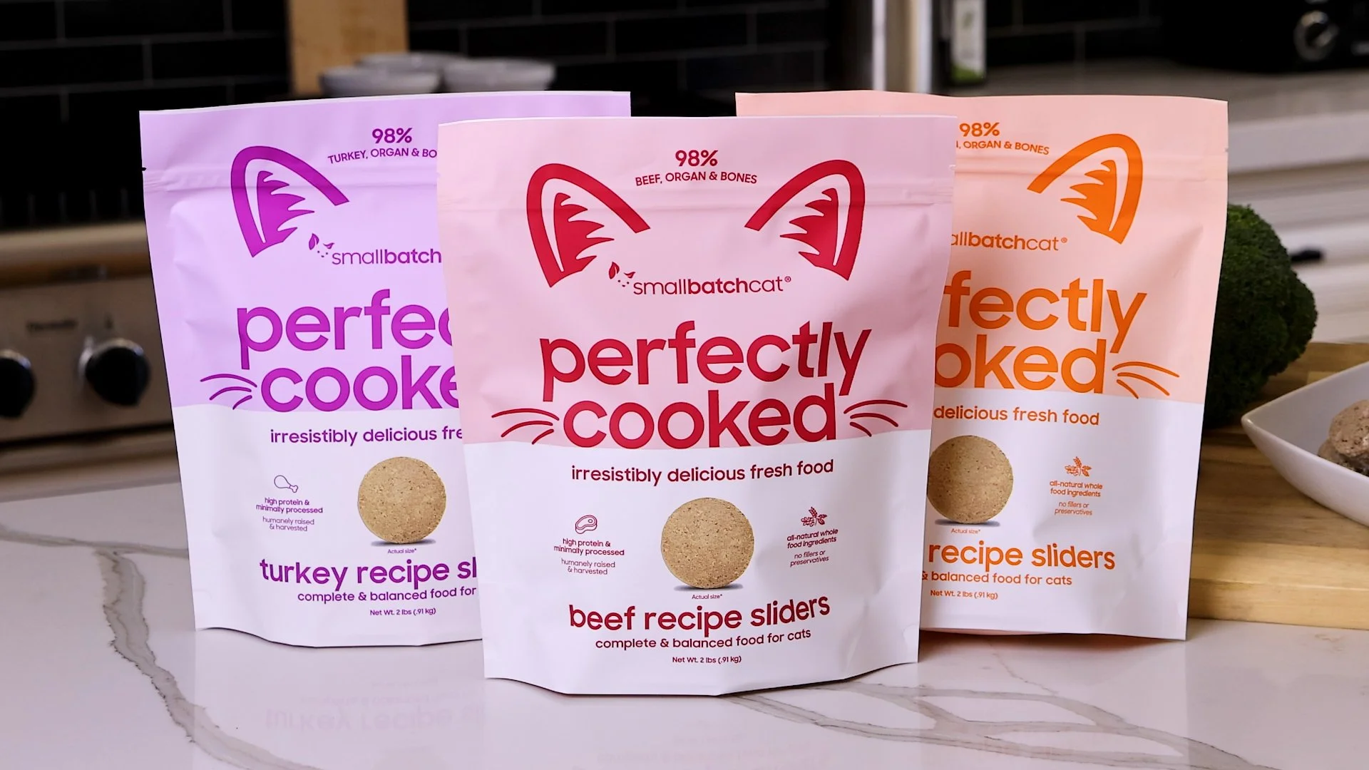 Three bags of smallbatchcat freeze-dried cat food on a white marble countertop. The bags are labeled 'perfectly cooked' with flavors 'turkey', 'beef', and 'fish,' and feature cat face or ear illustrations in purple, red, and orange, respectively.