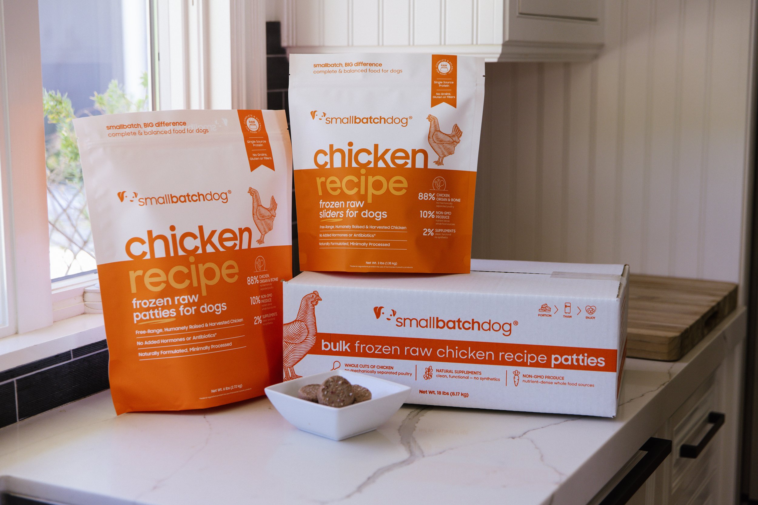 Boxes of smallbatchdog frozen raw dog food chicken patties and sliders on a kitchen countertop beside a small bowl with raw chicken patties with a window and white cabinets in the background.