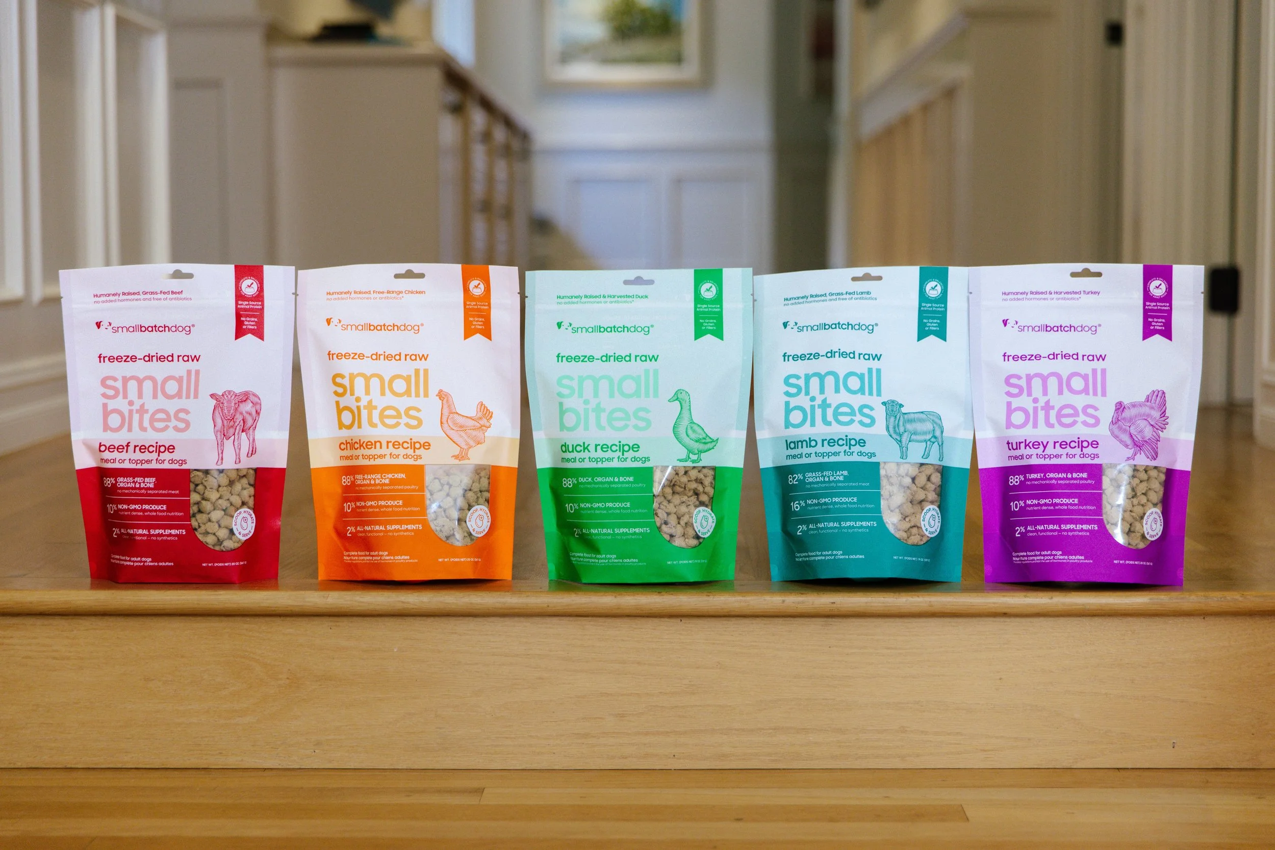 Five colorful bags of Smallbatchdog freeze-dried raw dog food lined up on a wooden surface.