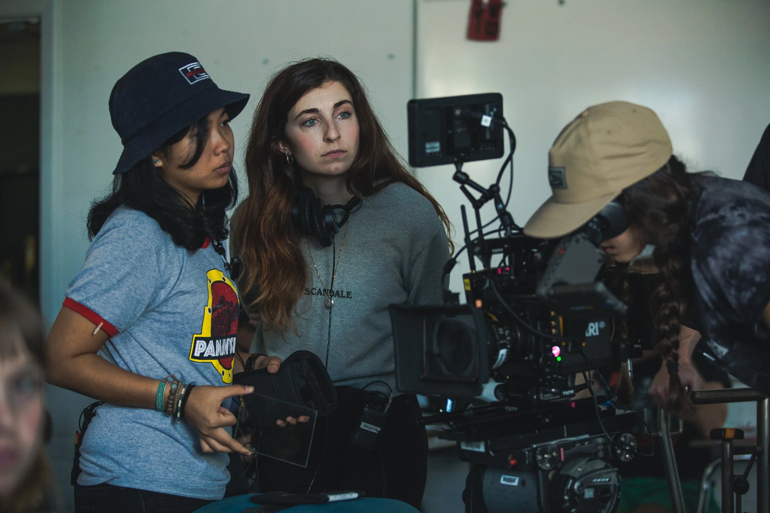 Group of women on a film set working with a professional camera, one woman in a gray sweatshirt and headphones looks directly at the camera, another woman with dark hair and a bucket hat adjusts the equipment.