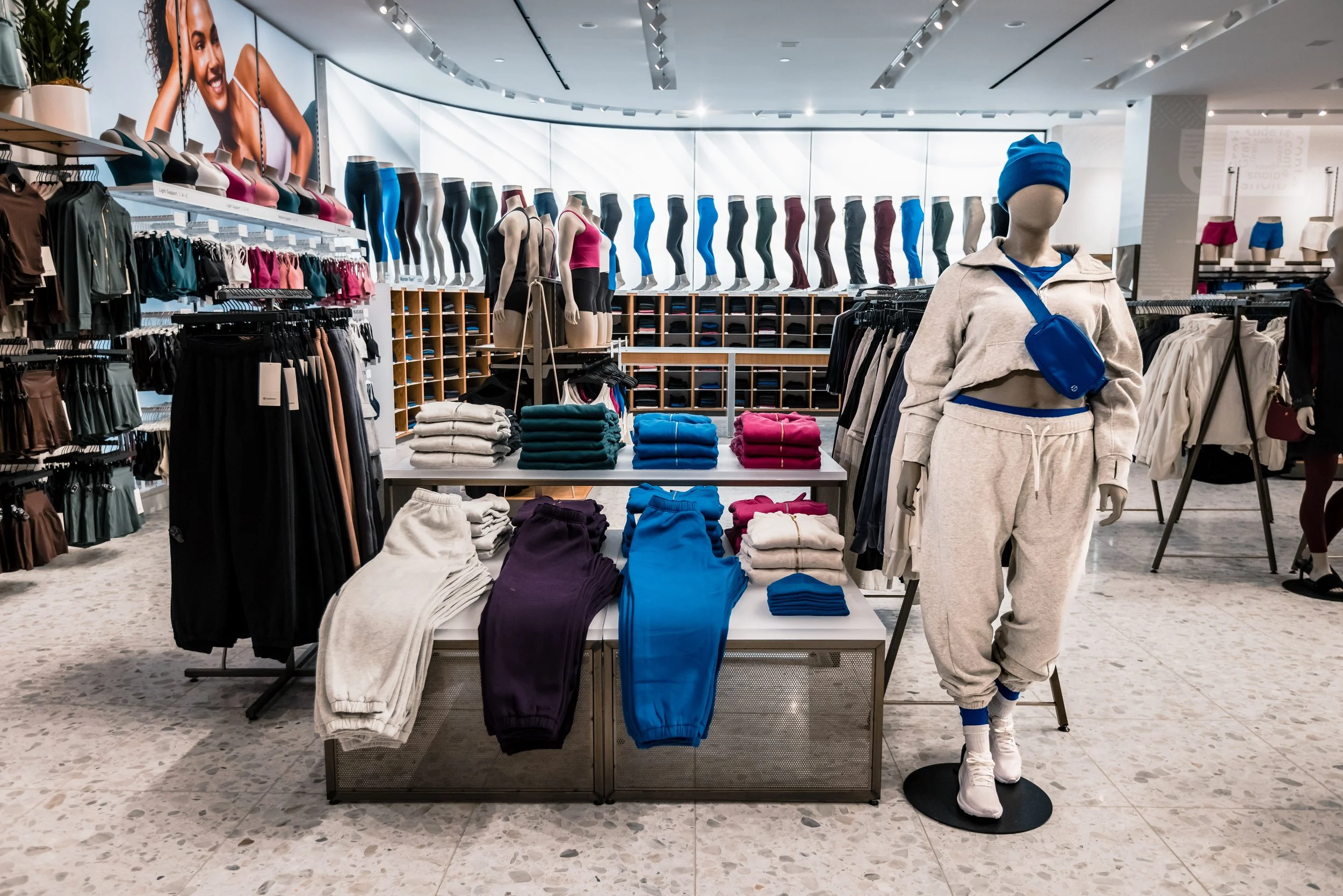 Clothing store display with mannequins dressed in athletic wear, folded sweatpants, t-shirts, and leggings in various colors, set with shelves of shoes in the background, and a large advertisement poster on the wall.