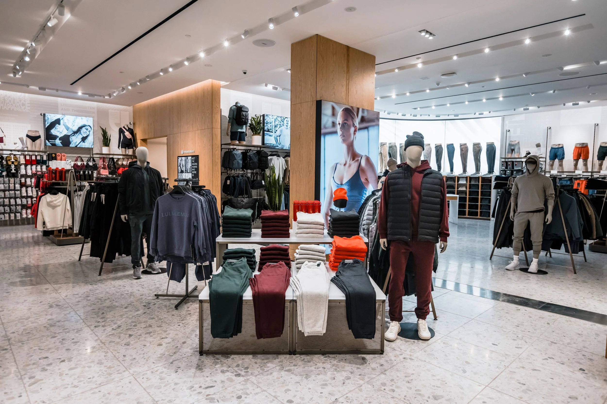 Inside a retail clothing store with mannequins dressed in athletic and casual wear, shelves of folded clothes, and digital screens showing models.