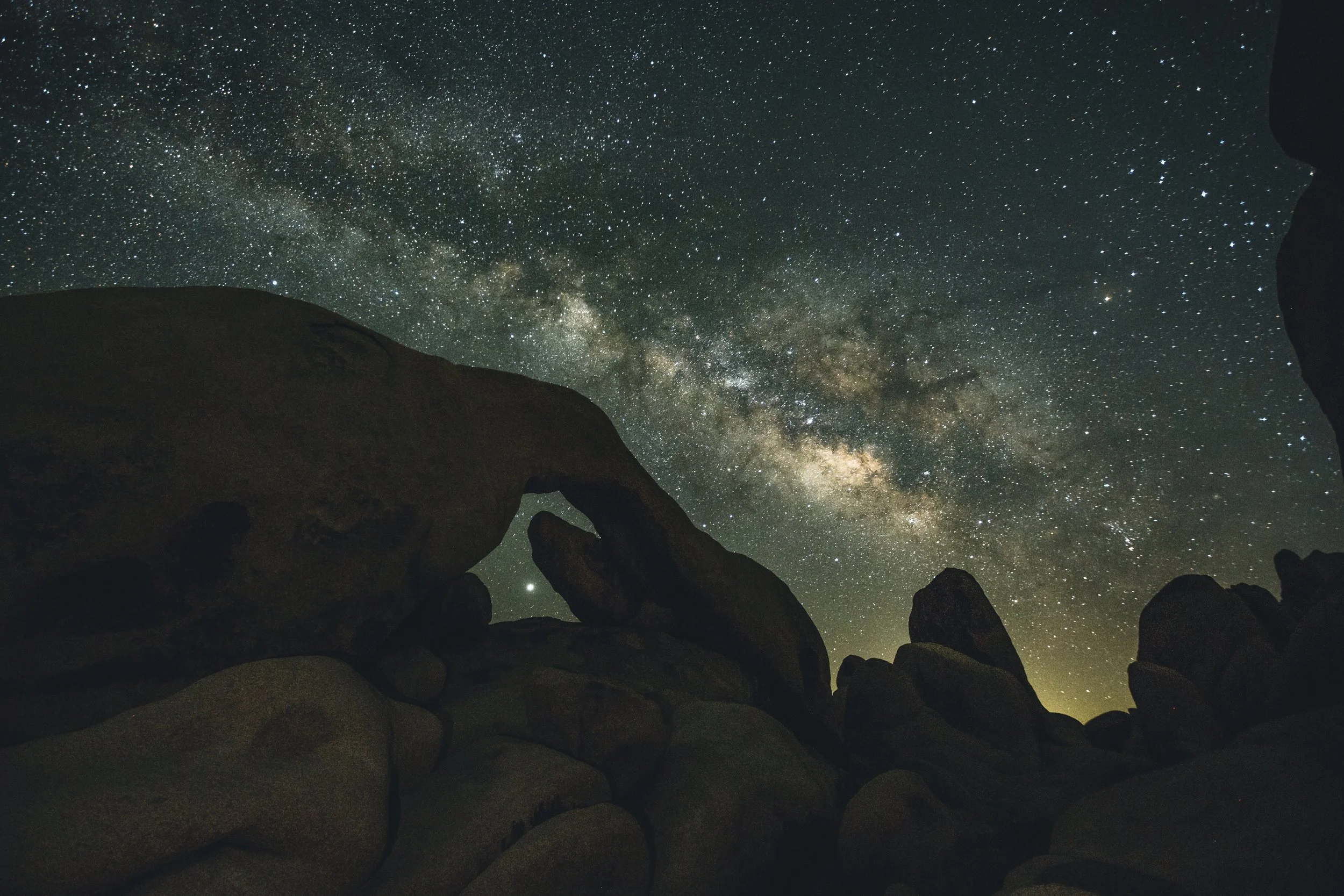 Night sky filled with stars and the Milky Way galaxy over dark rocky landscape with arch-shaped rock formation.