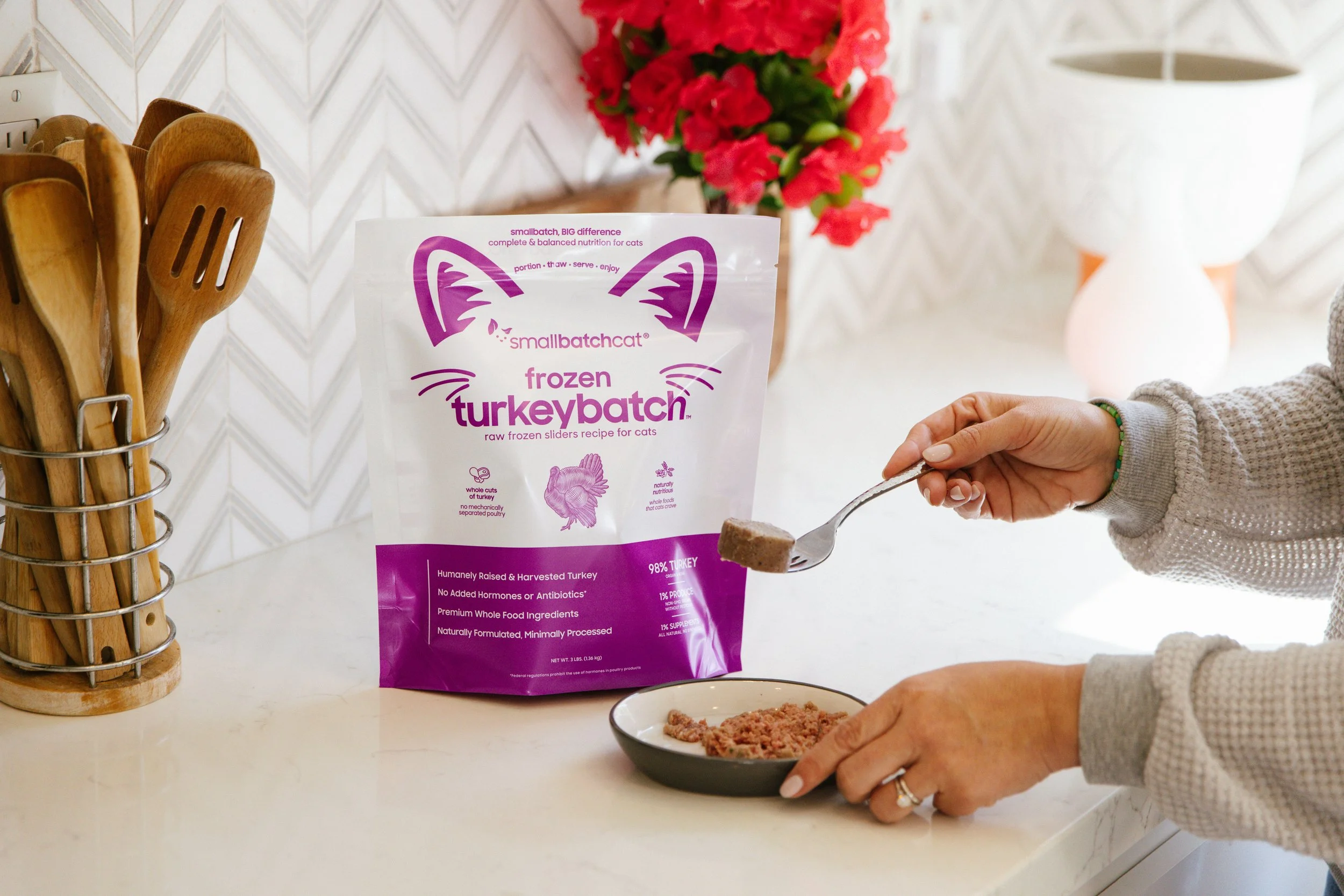 Person spooning frozen turkeycat cat food into a bowl in a kitchen, with a bag of Smallbatchcat frozen turkeybat ch cat food on the countertop, a container of wooden utensils in a holder, pink flowers in the background, and a white lamp on the side.