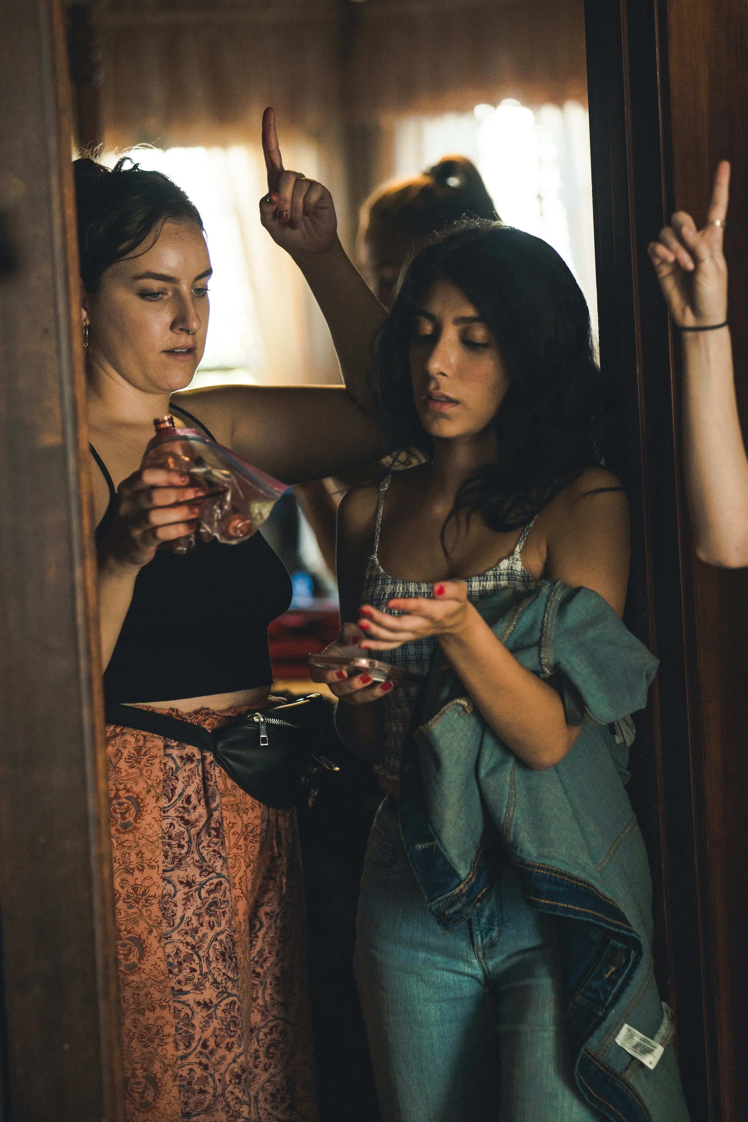 Two women in an elevator, one is holding a bottle of liquor and the other a smartphone, with another woman partially visible behind them making a peace sign.