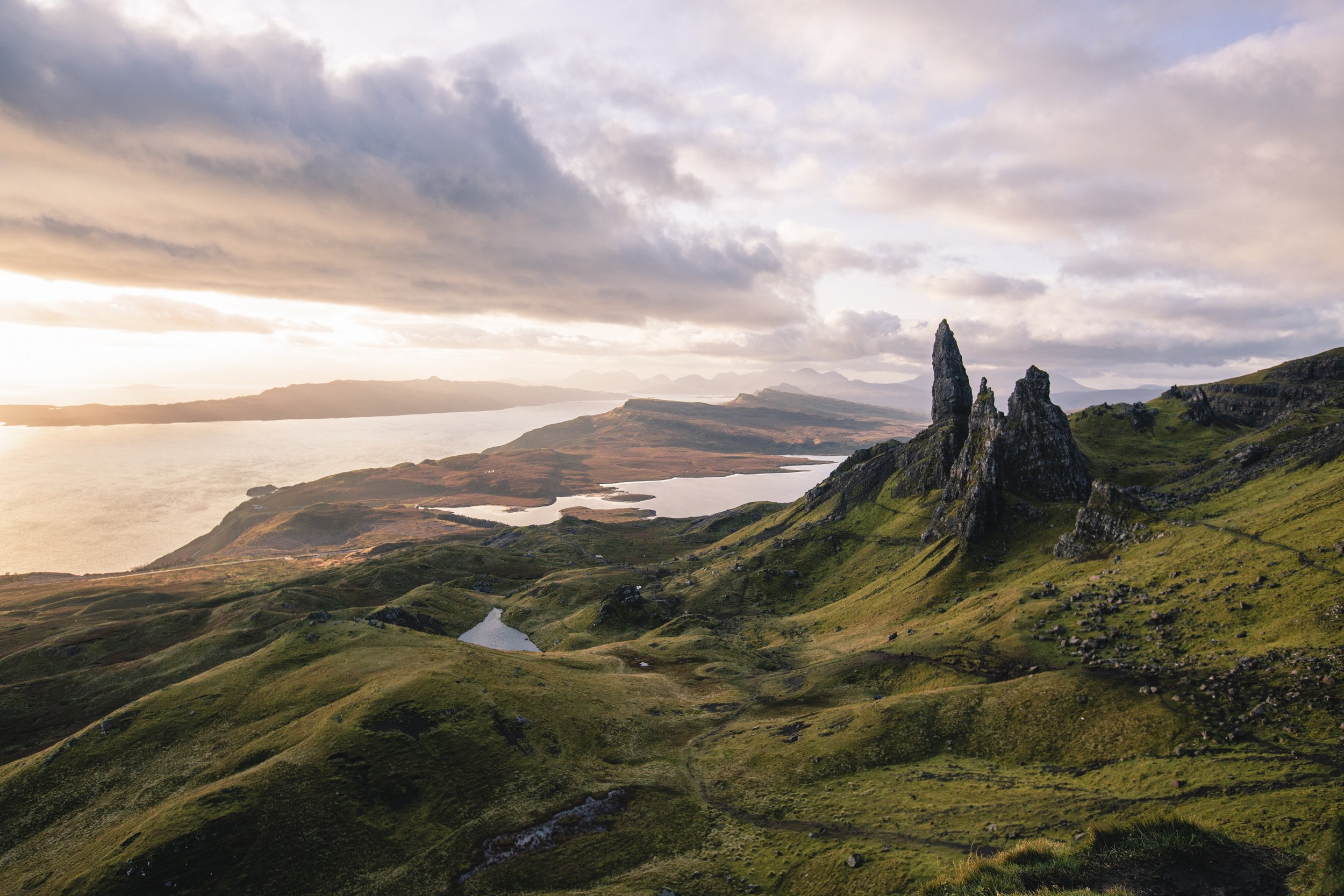 Scenic view of green rolling hills, rocky outcroppings, lakes, and a distant body of water under a cloudy sky at sunset or sunrise.