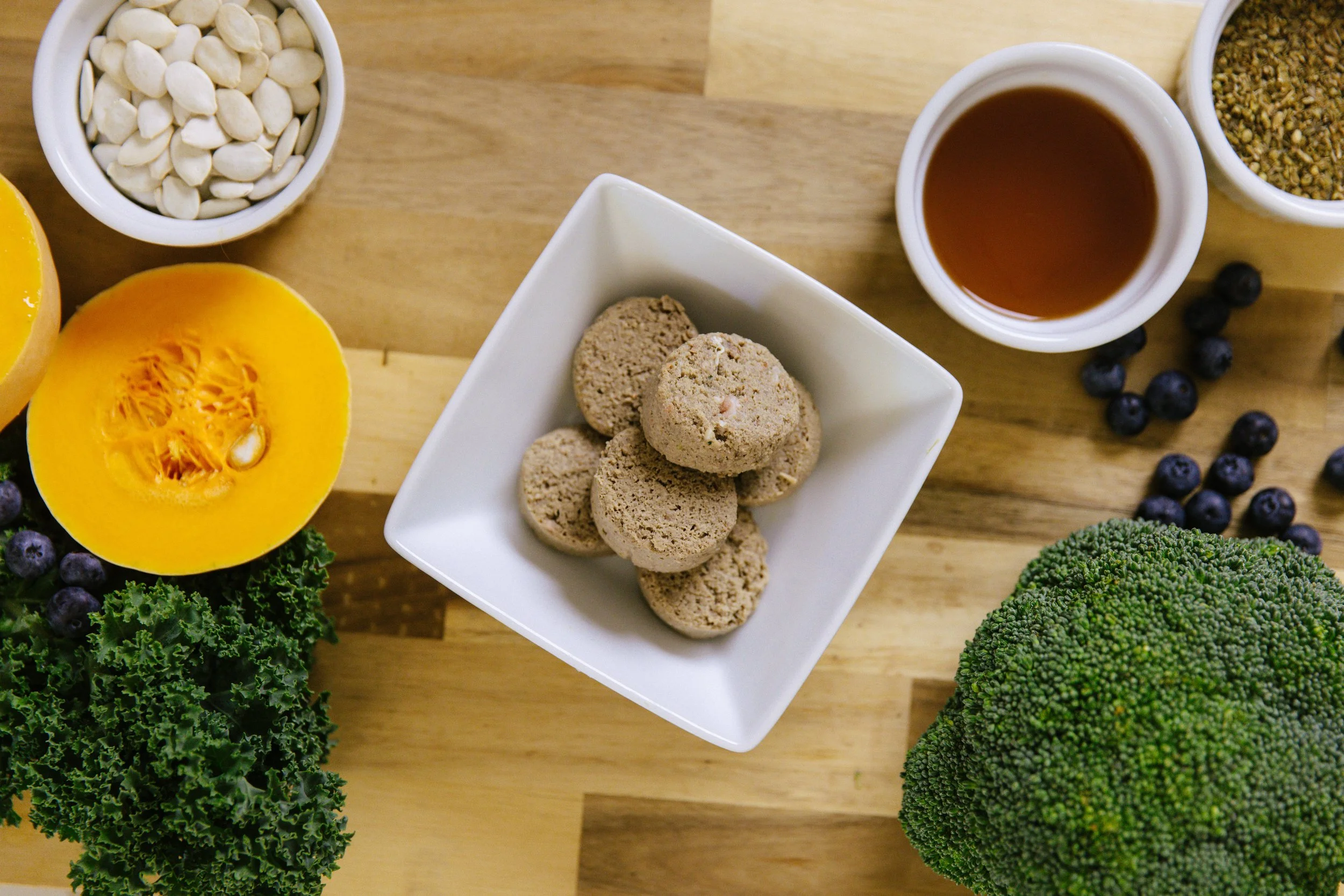 A wooden table with a halved yellow pumpkin, a bowl of white pumpkin seeds, a small white bowl of brown liquid syrup, a small bowl of green herbs or spices, a cluster of blueberries, and a white square bowl filled with round beige cookies or biscuits