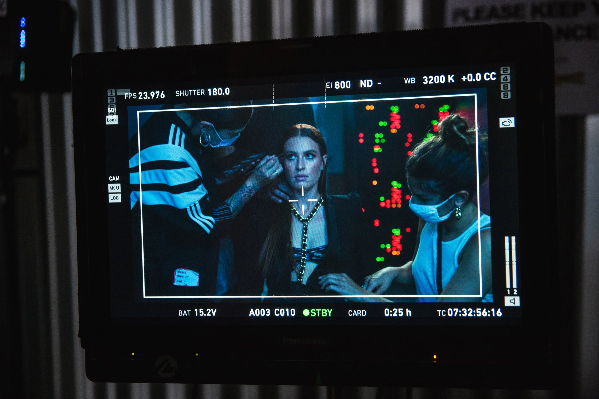 A behind-the-scenes shot showing a woman getting makeup applied by a makeup artist, with another person assisting, through a camera monitor on a film set.