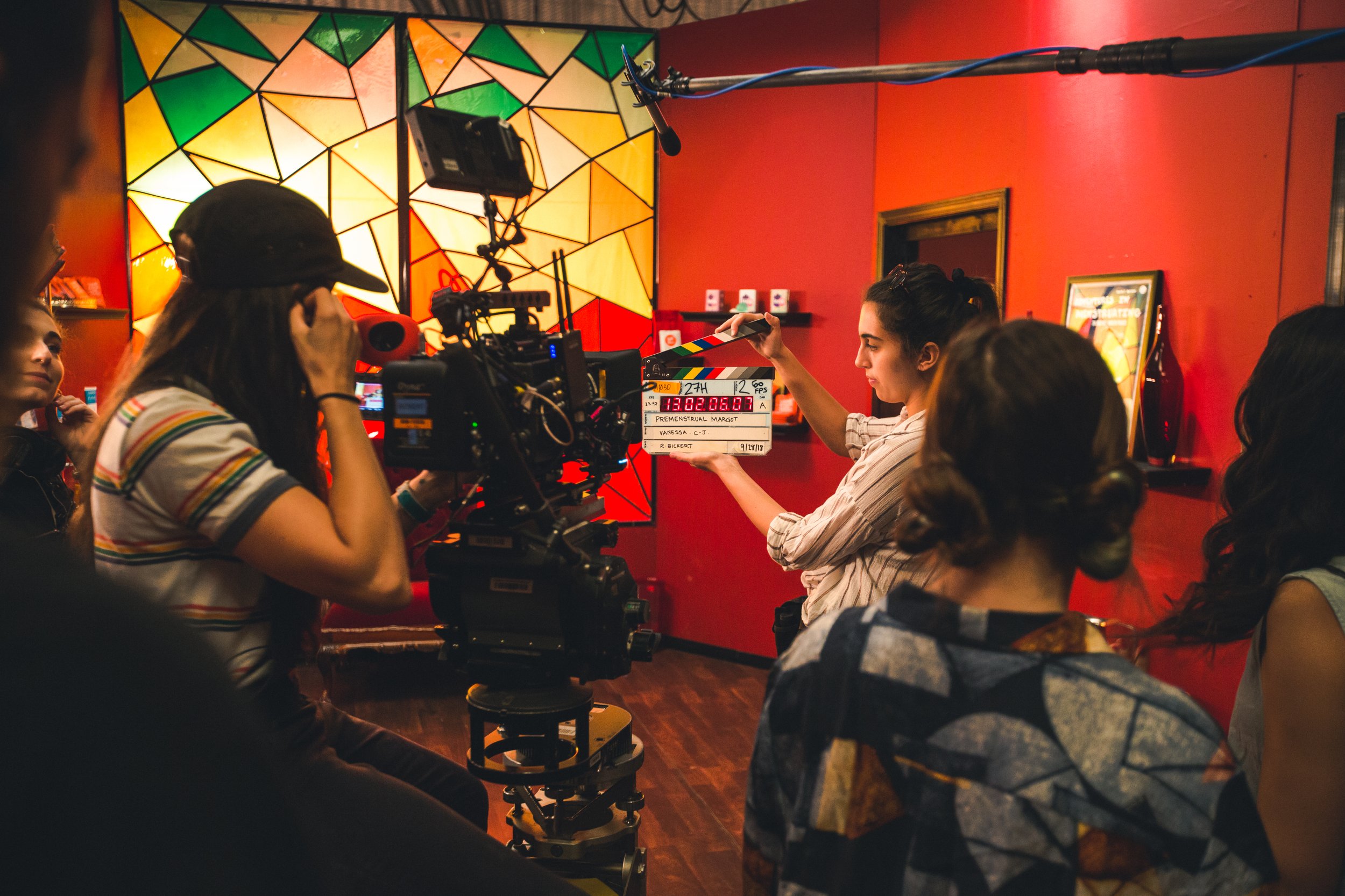 Film crew filming a woman holding a clapperboard inside a room with red walls and colorful stained glass artwork.