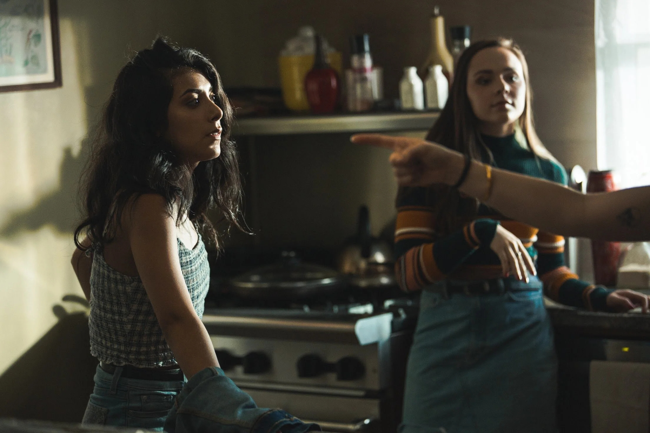 Two young women in a kitchen, one with dark, wavy hair and patterned sleeveless top looking to the side, the other with long straight hair and striped sweater looking at the camera, while a third person points with their index finger toward the woman