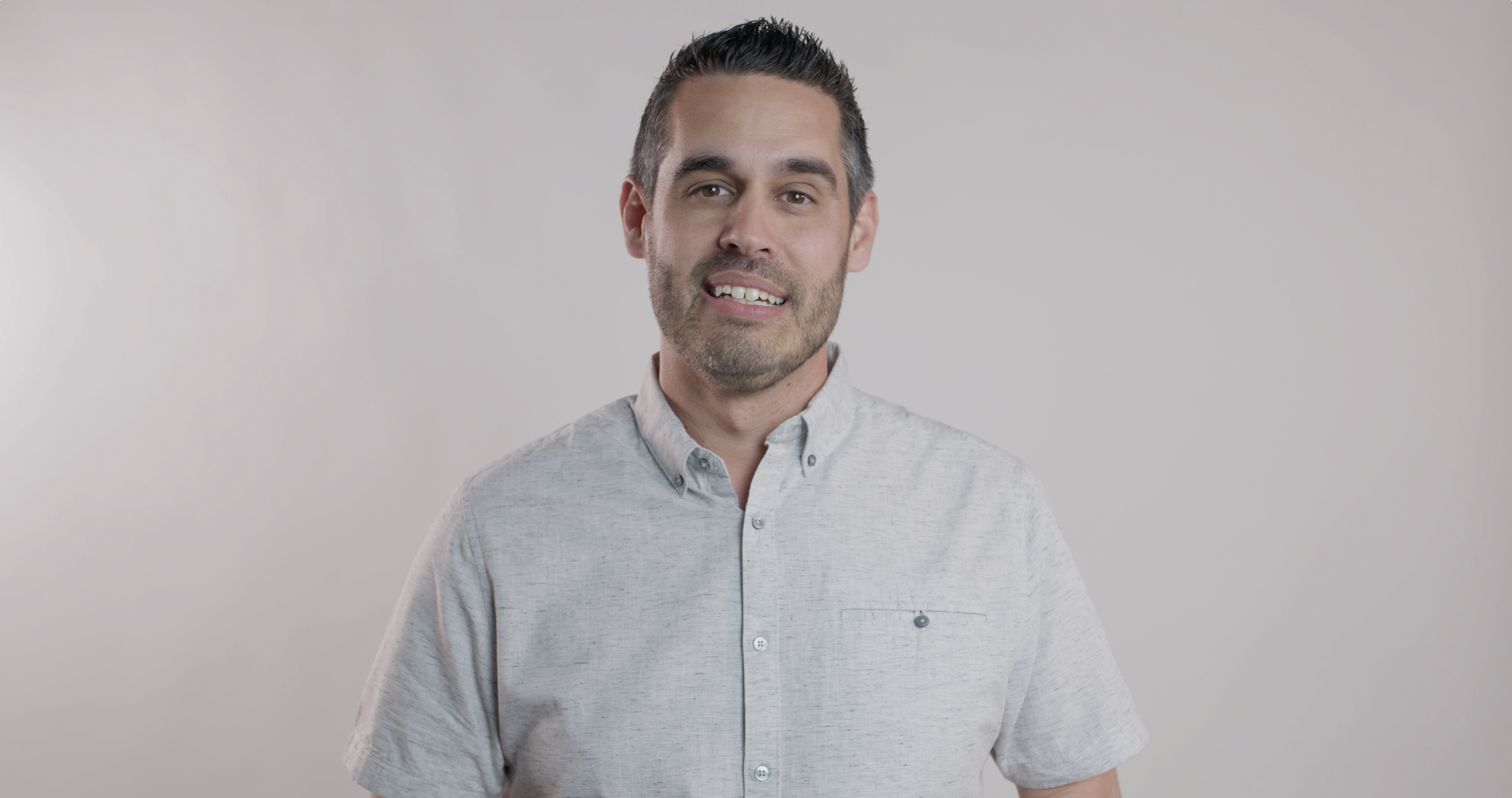 Headshot of a man with short dark hair, slight beard, wearing a light gray button-up shirt, standing against a plain white background.