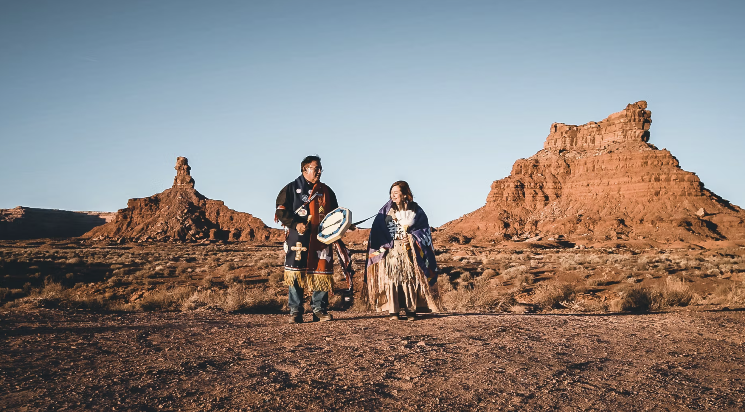 Two Indigenous individuals in traditional clothing standing in a desert landscape with large rock formations in the background.