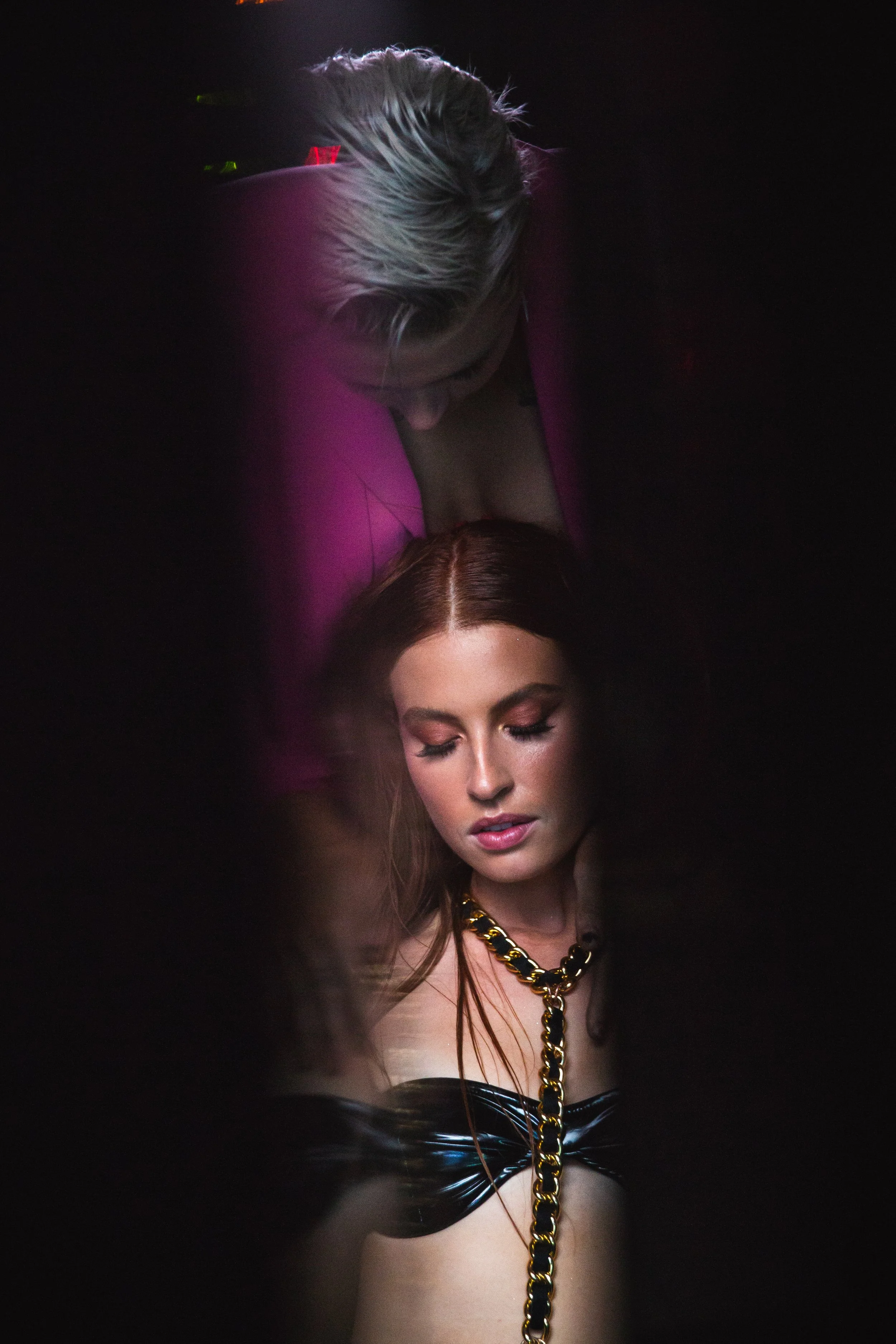 A woman with long red hair and makeup, wearing a black strapless top with a large gold and black chain necklace, is looking downward with her eyes closed. Behind her, a woman with blonde hair and a purple jacket is looking down at her from above.