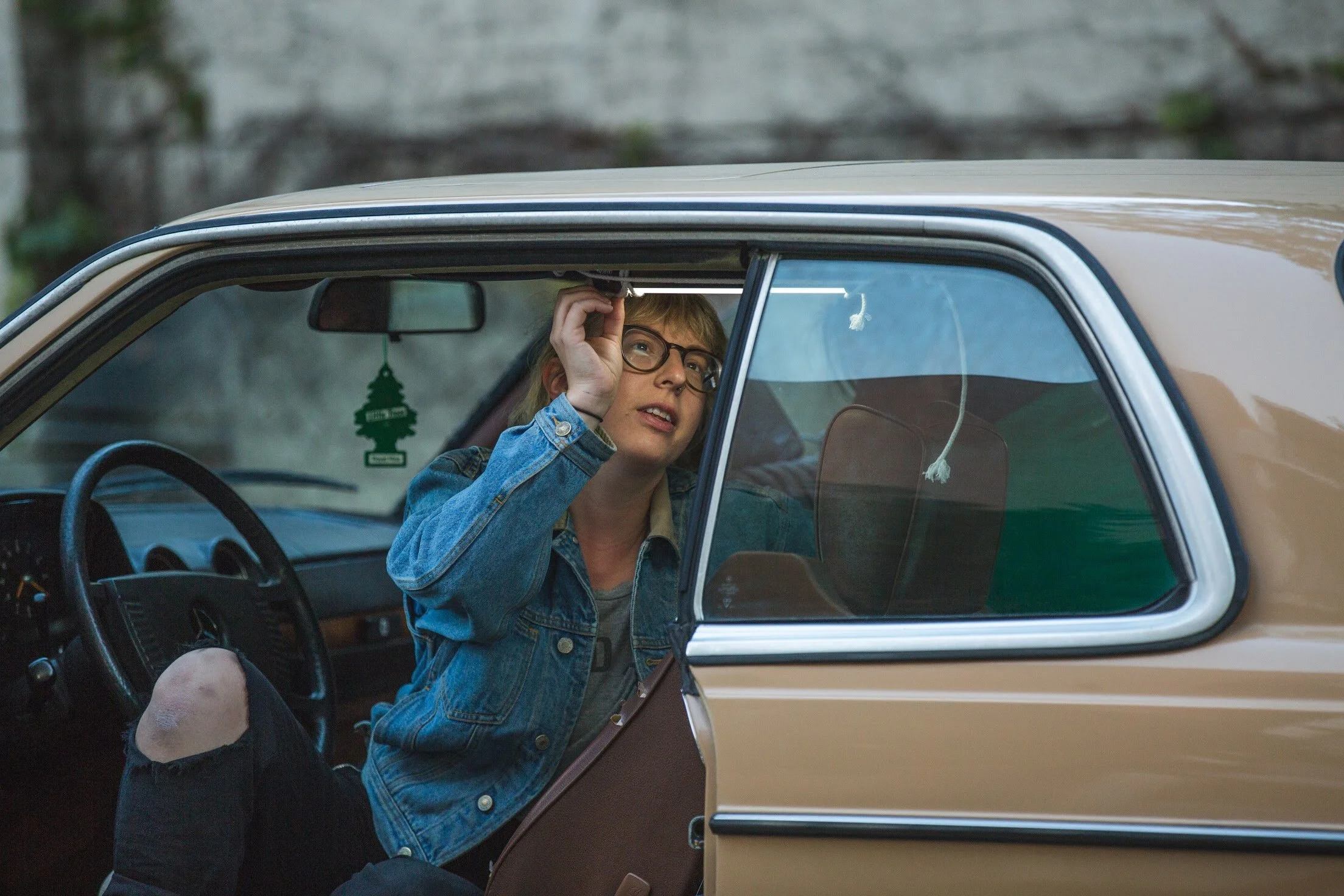 Woman with glasses, blonde hair, wearing a denim jacket, sitting inside a vintage beige car, adjusting her glasses and looking out the window.