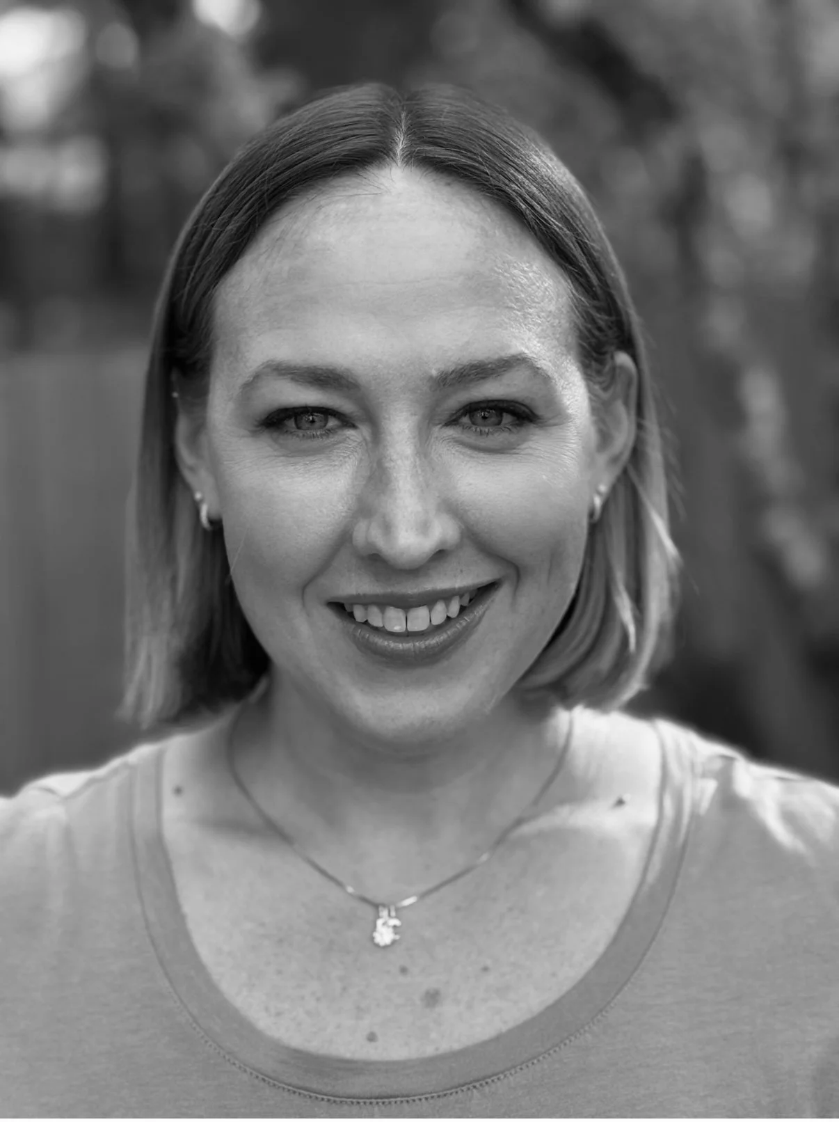 A smiling woman with shoulder-length hair takes a selfie outdoors, with trees and a fence in the background.