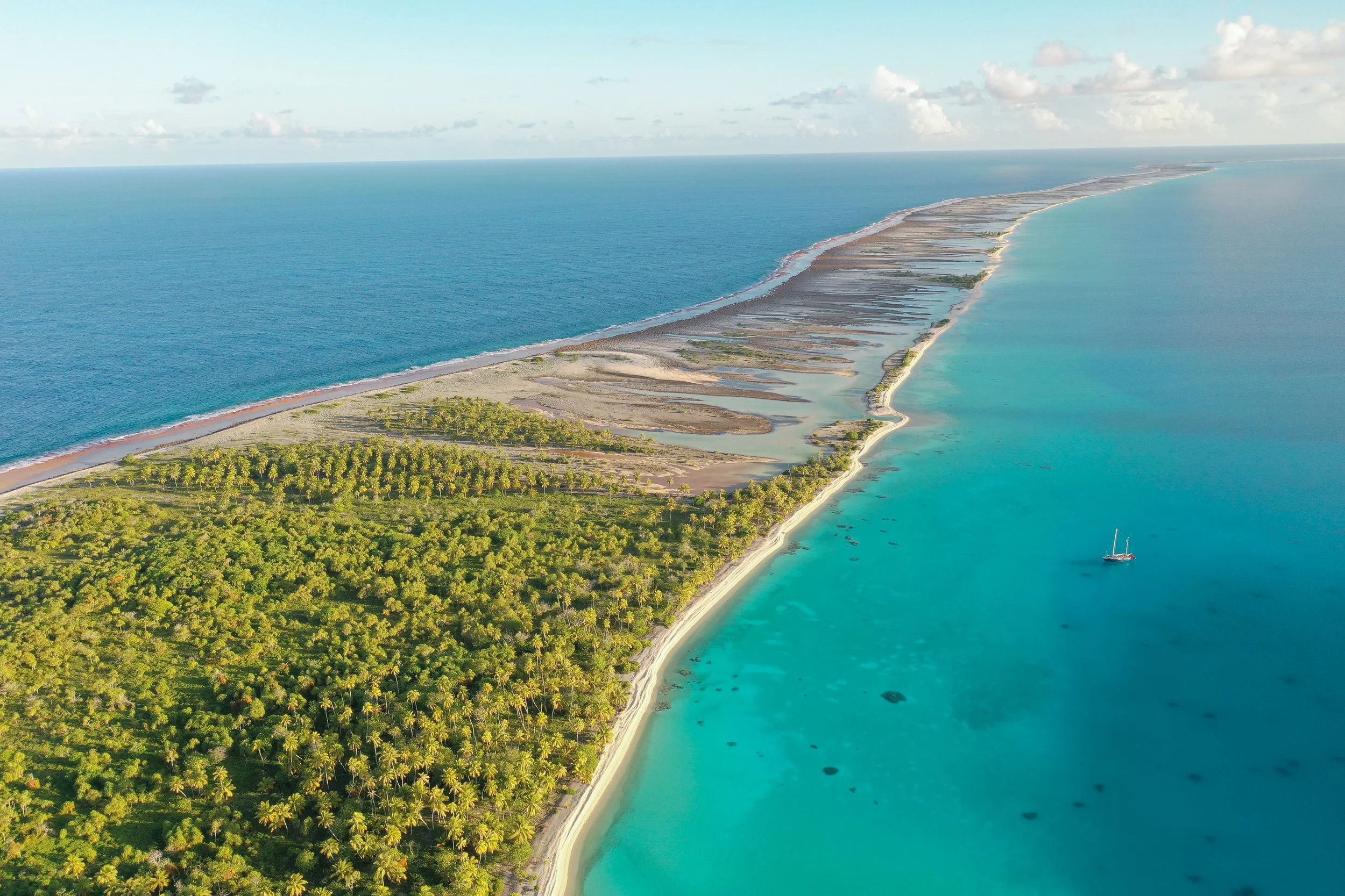 Aerial view of a tropical beach with green vegetation on one side and turquoise waters on the other, with a sailboat anchored near the shoreline.