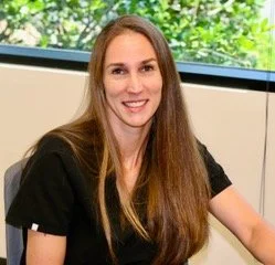 A woman with long brown hair, wearing a black top, sitting at a desk in an office environment with a window showing trees in the background.