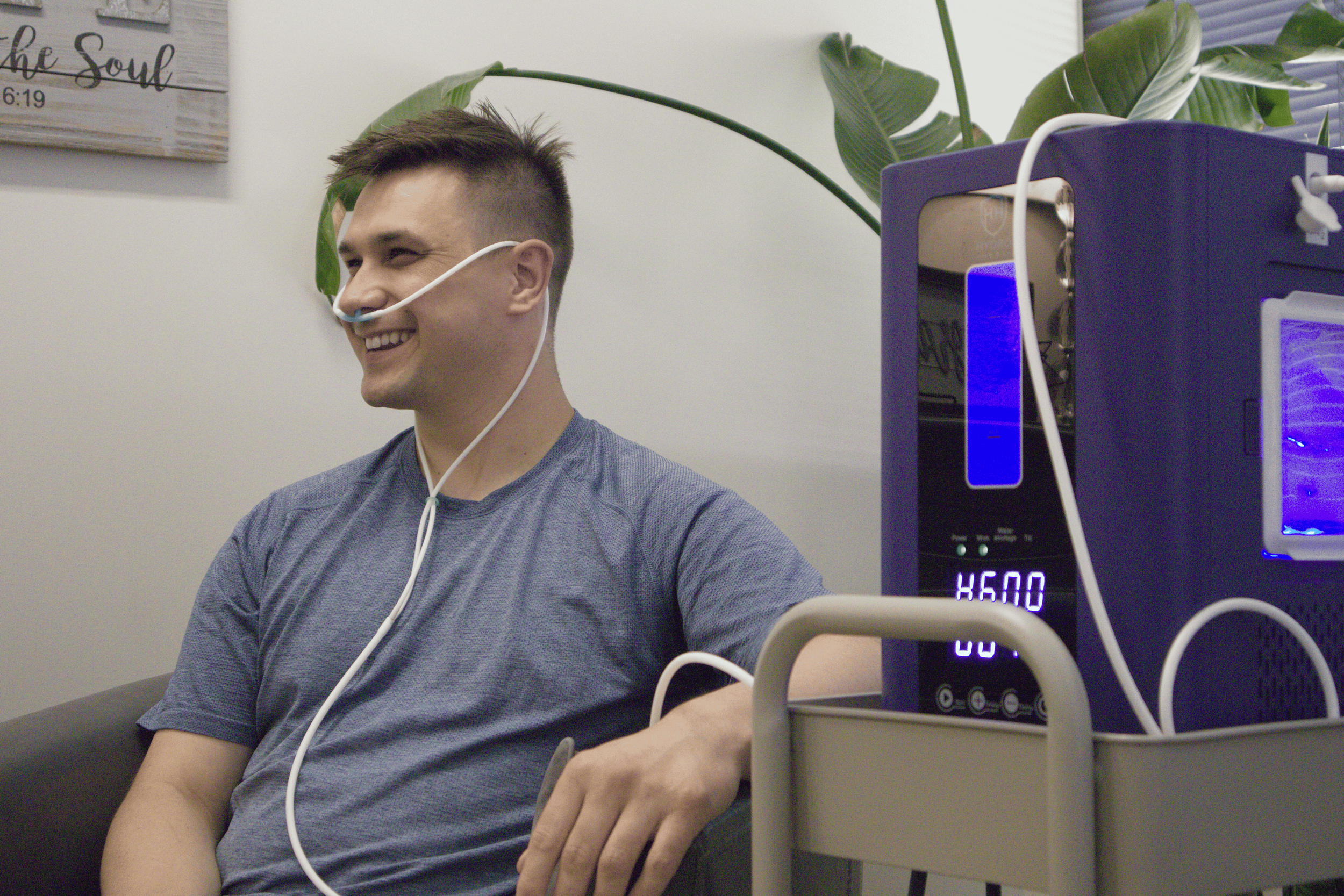 A young man smiling while receiving hydroge therapy with a nasal tube. There is a large green plant and a decorative sign on the wall that reads 'the soul' in the background.
