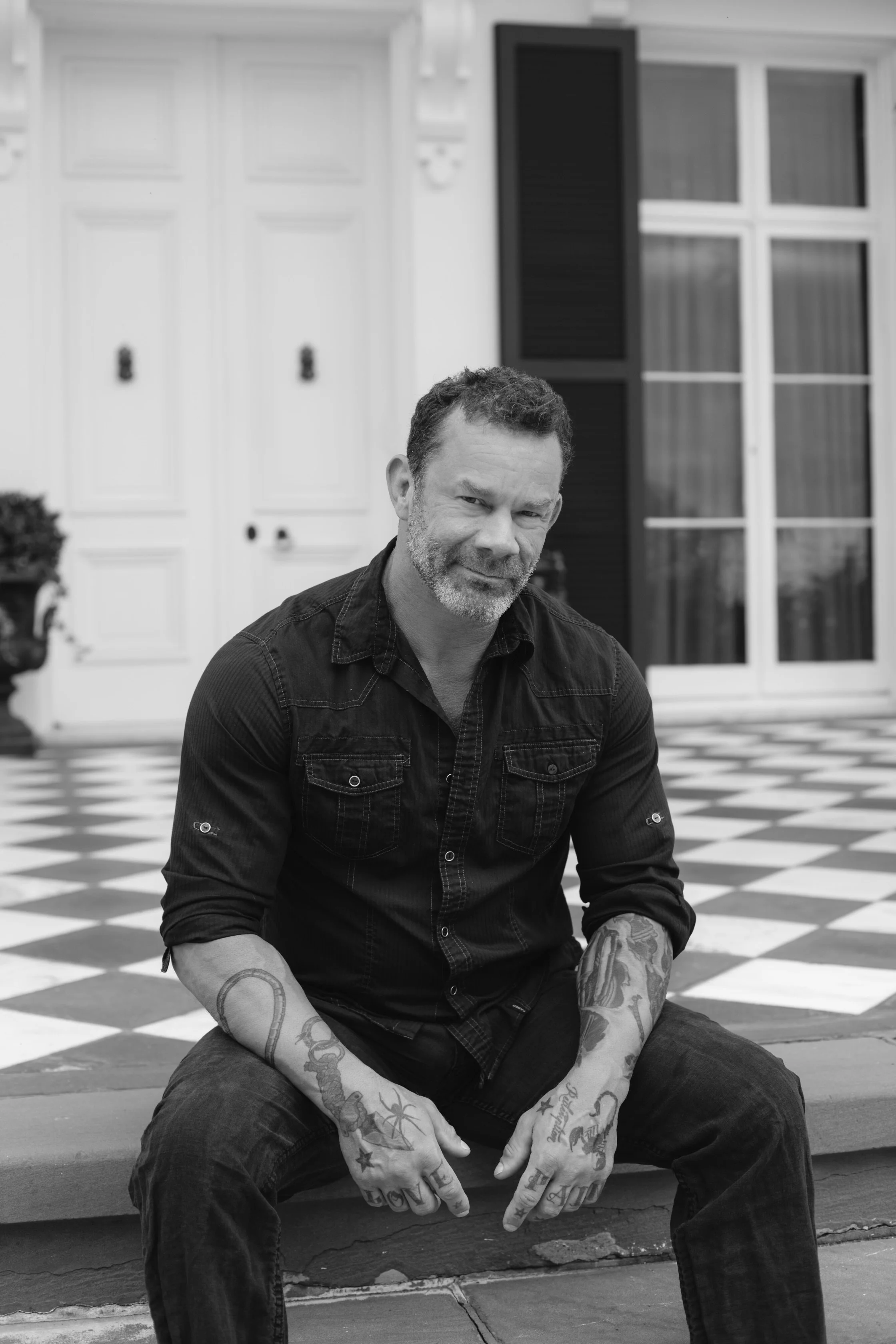 A man with tattoos sitting in front of a house, with a checkered black and white floor and a white door.