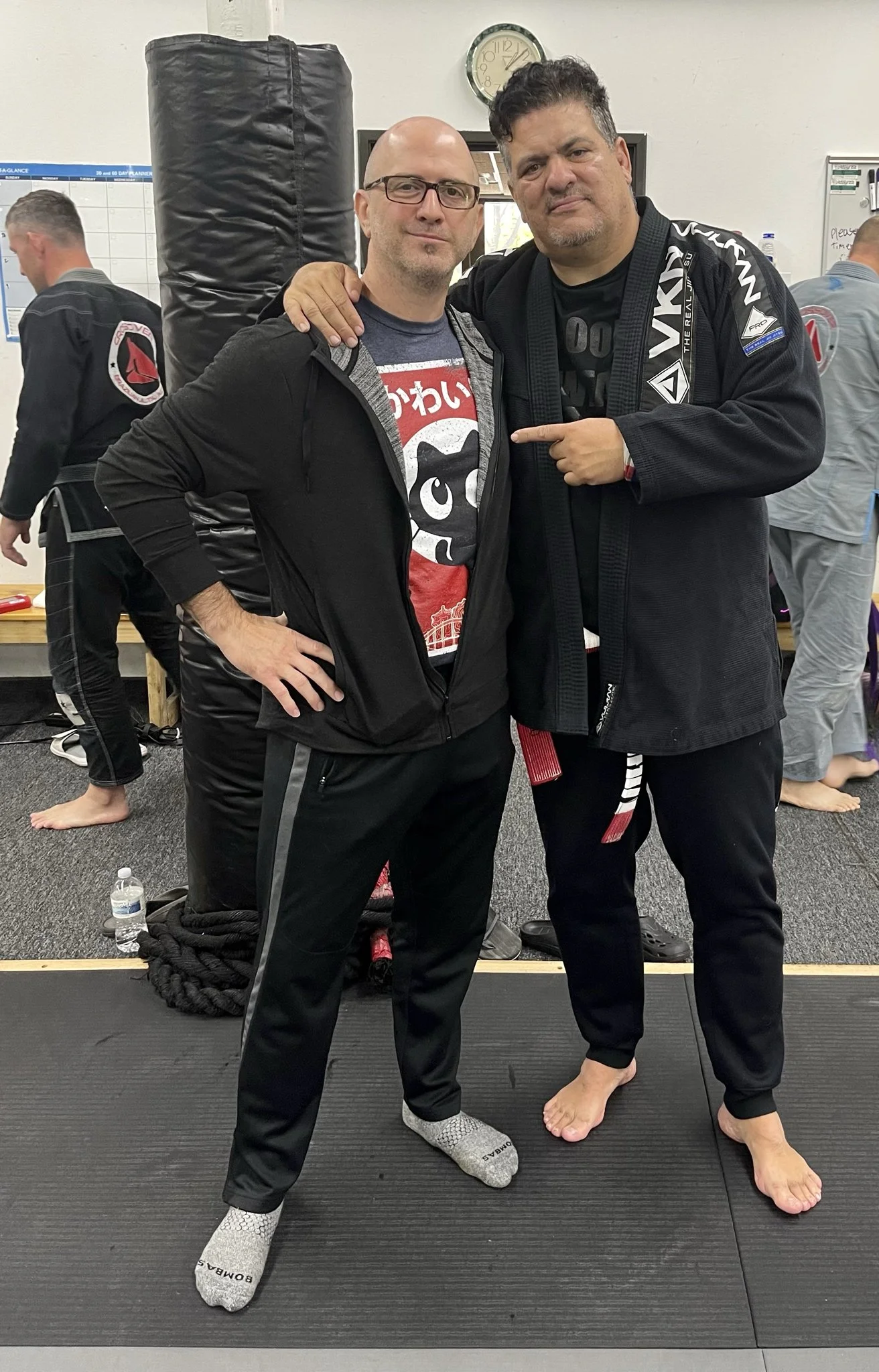 Two men in Brazilian Jiu-Jitsu gis standing on a mat in a gym, posing for a photo with two other men training in the background. One man has his arm around the other's shoulder, and the other man is pointing at the first. One man is wearing socks, and the other is barefoot.