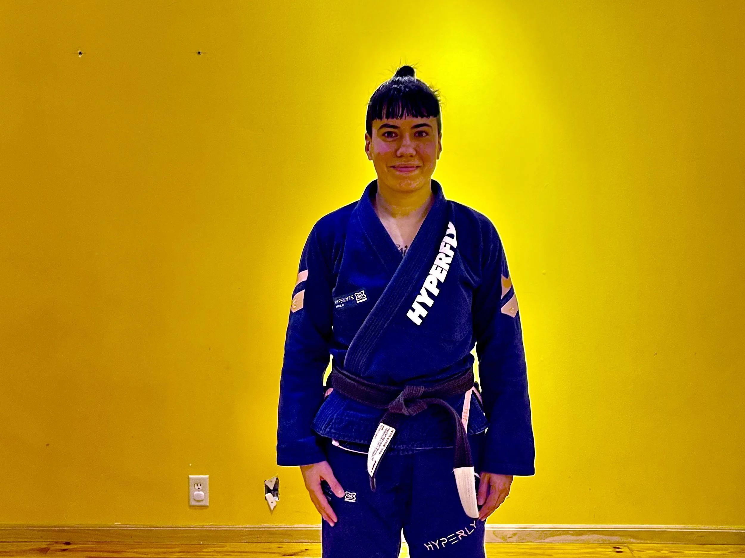 A woman in a blue martial arts gi with a black belt standing in a room with a yellow wall.