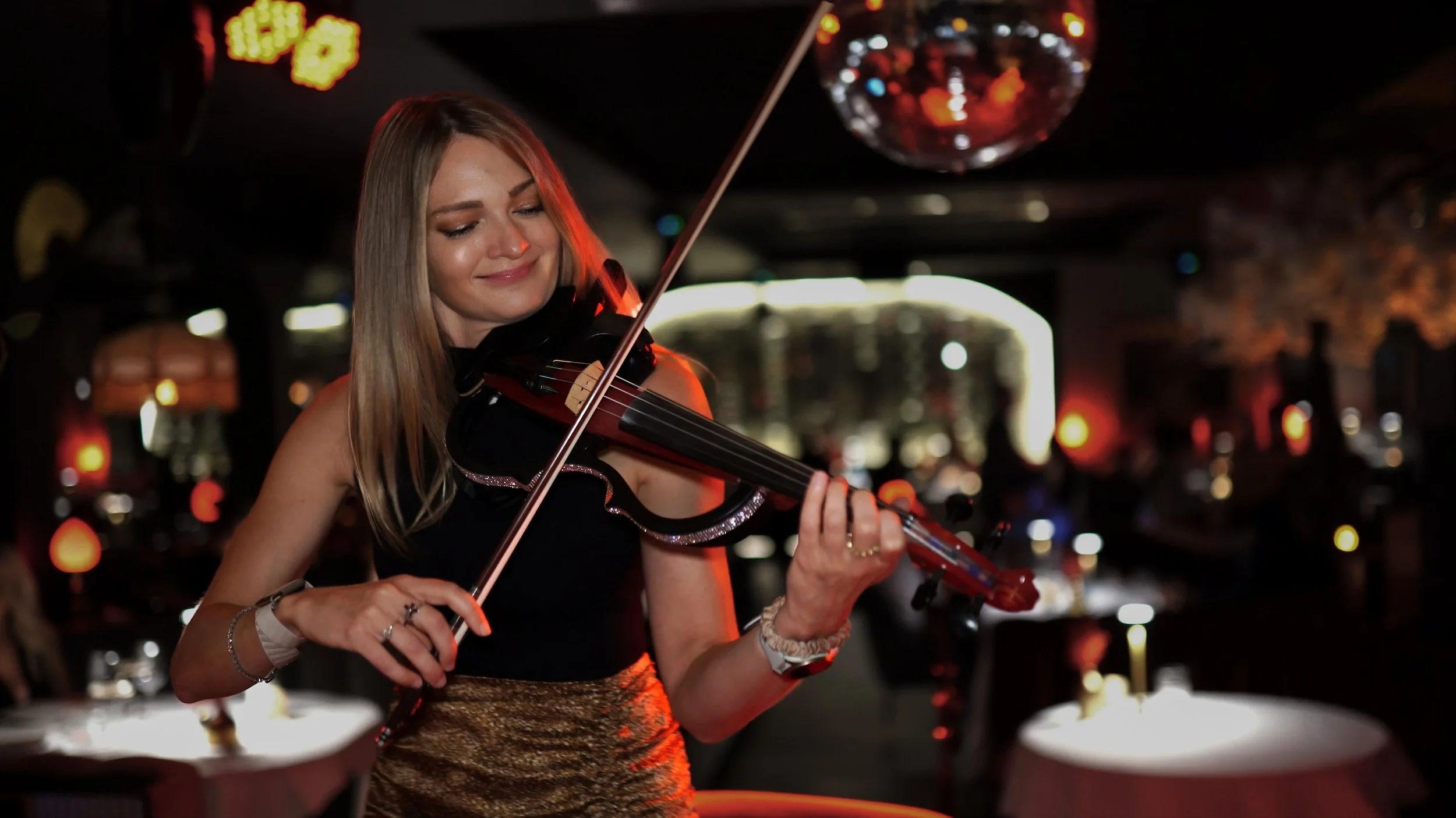 A woman playing a violin in a dimly lit restaurant or club with a disco ball overhead and blurred background.