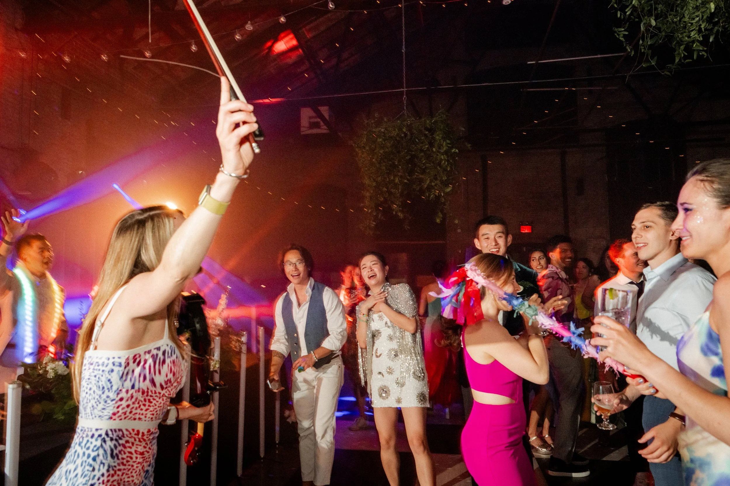 People celebrating at a party with a woman holding a flag, others wearing festive attire, smiling, and enjoying drinks in a lively indoor setting with colorful lighting.