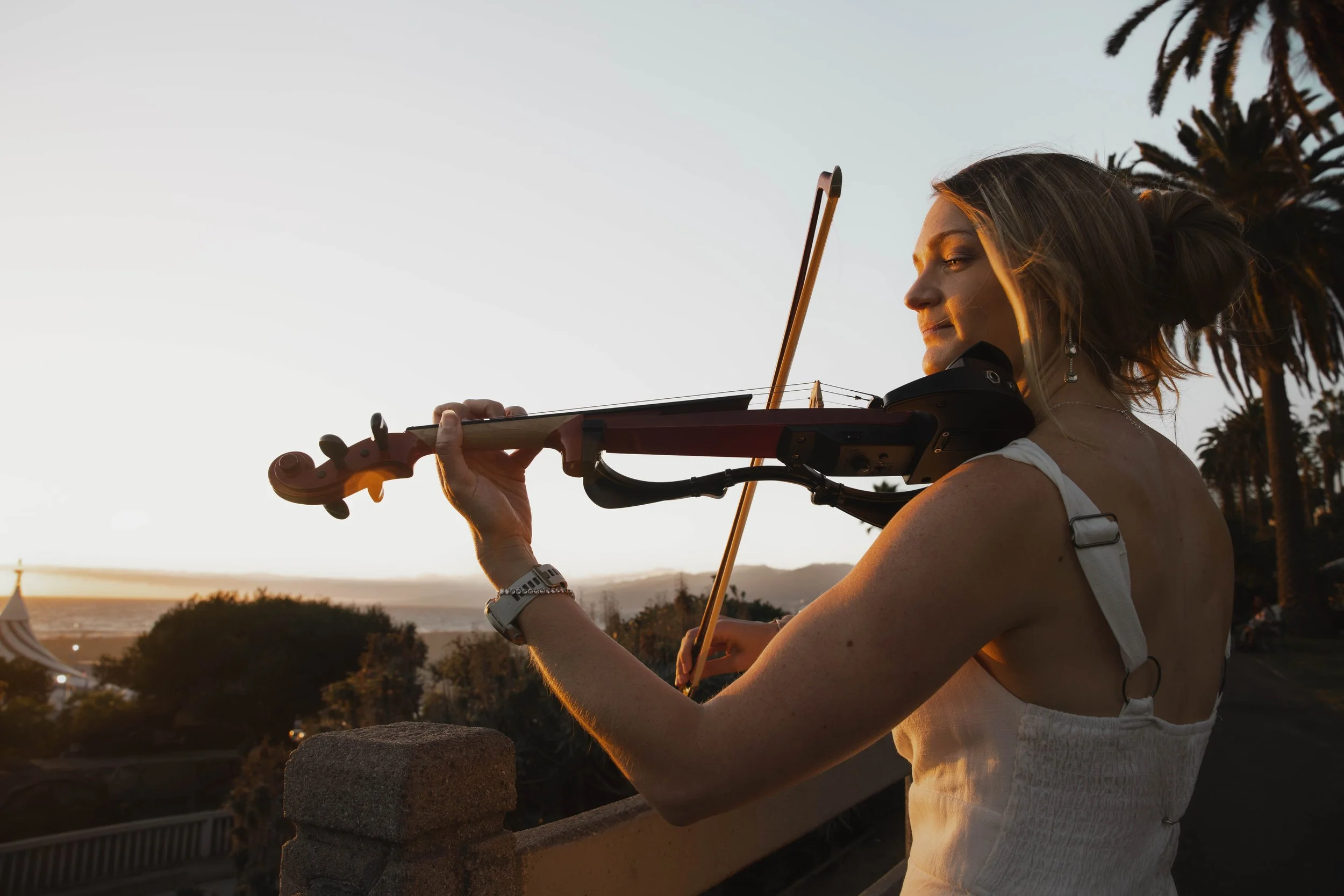 Woman playing violin outdoors during sunset, with palm trees in the background.