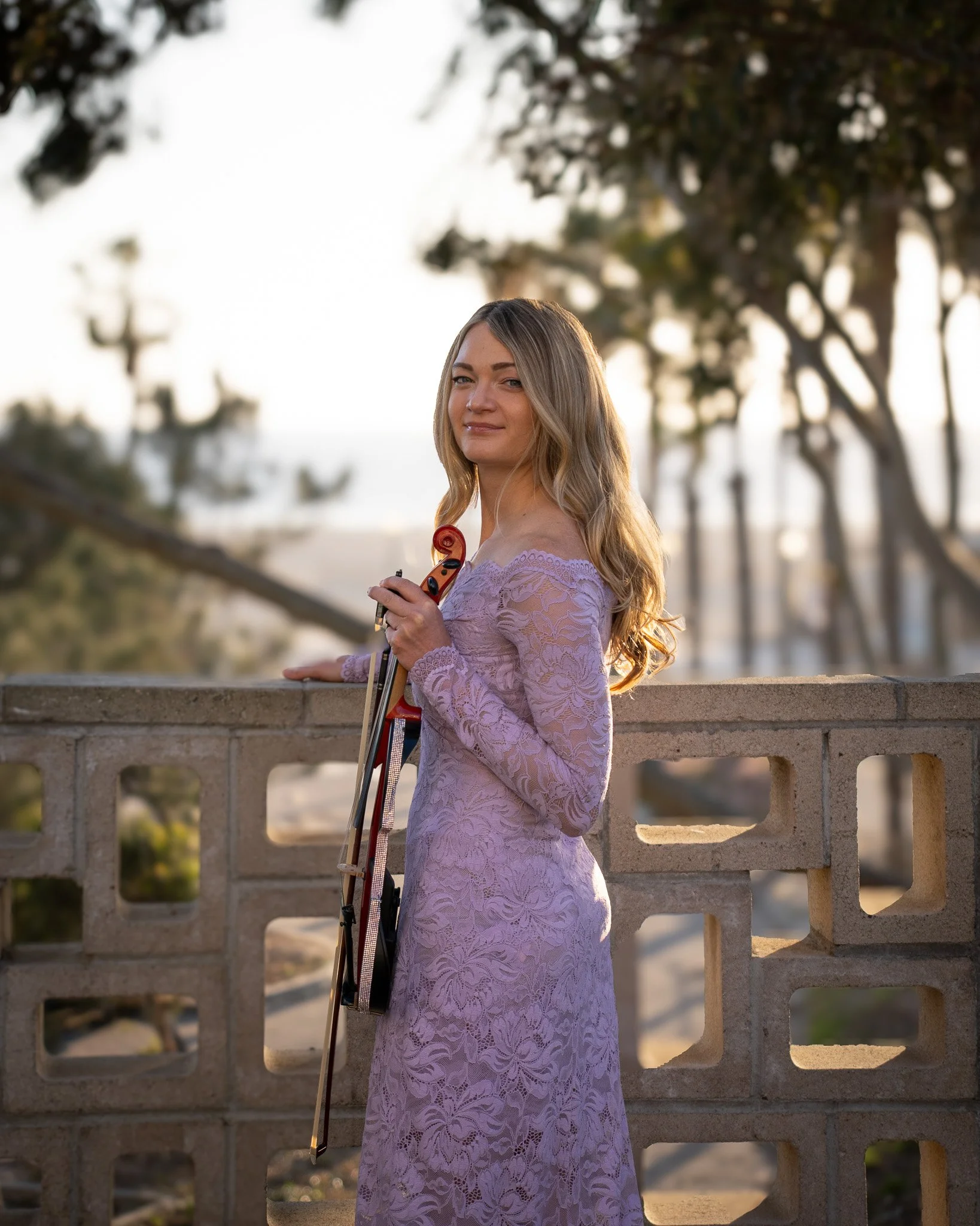 A woman in a lavender lace dress holding a violin and bow, standing outdoors near a brick wall with trees and sunlight in the background.