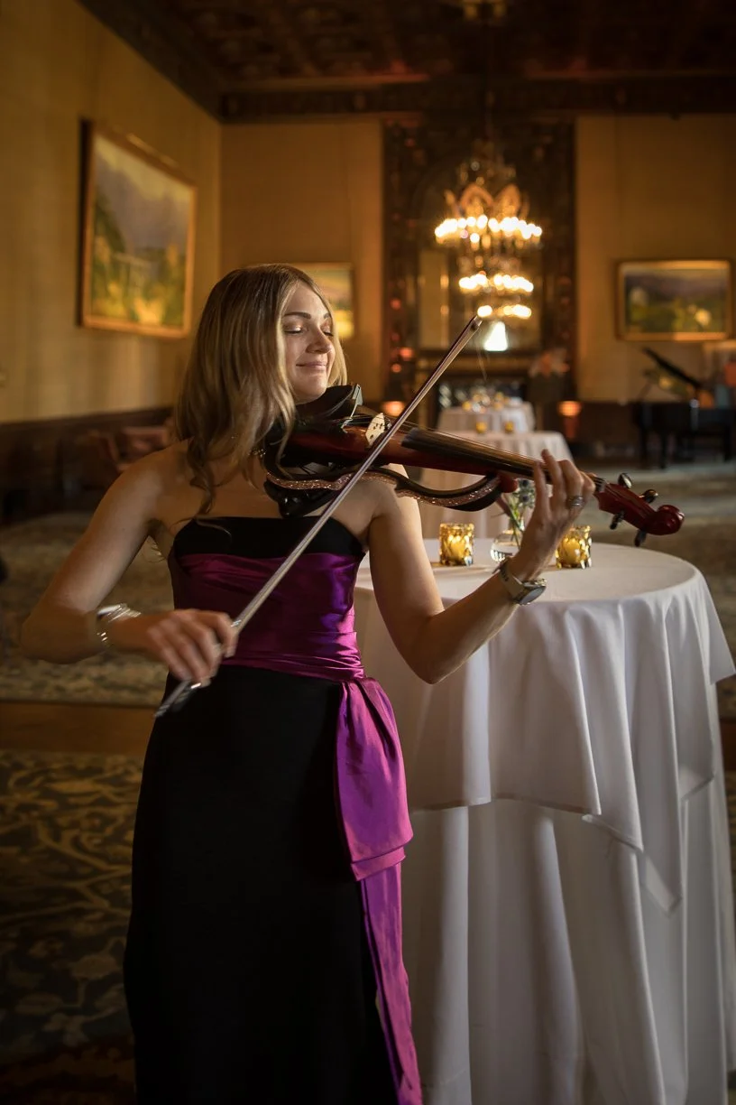 A woman playing the violin in an elegant, dimly lit room with paintings on the walls and a chandelier in the background.