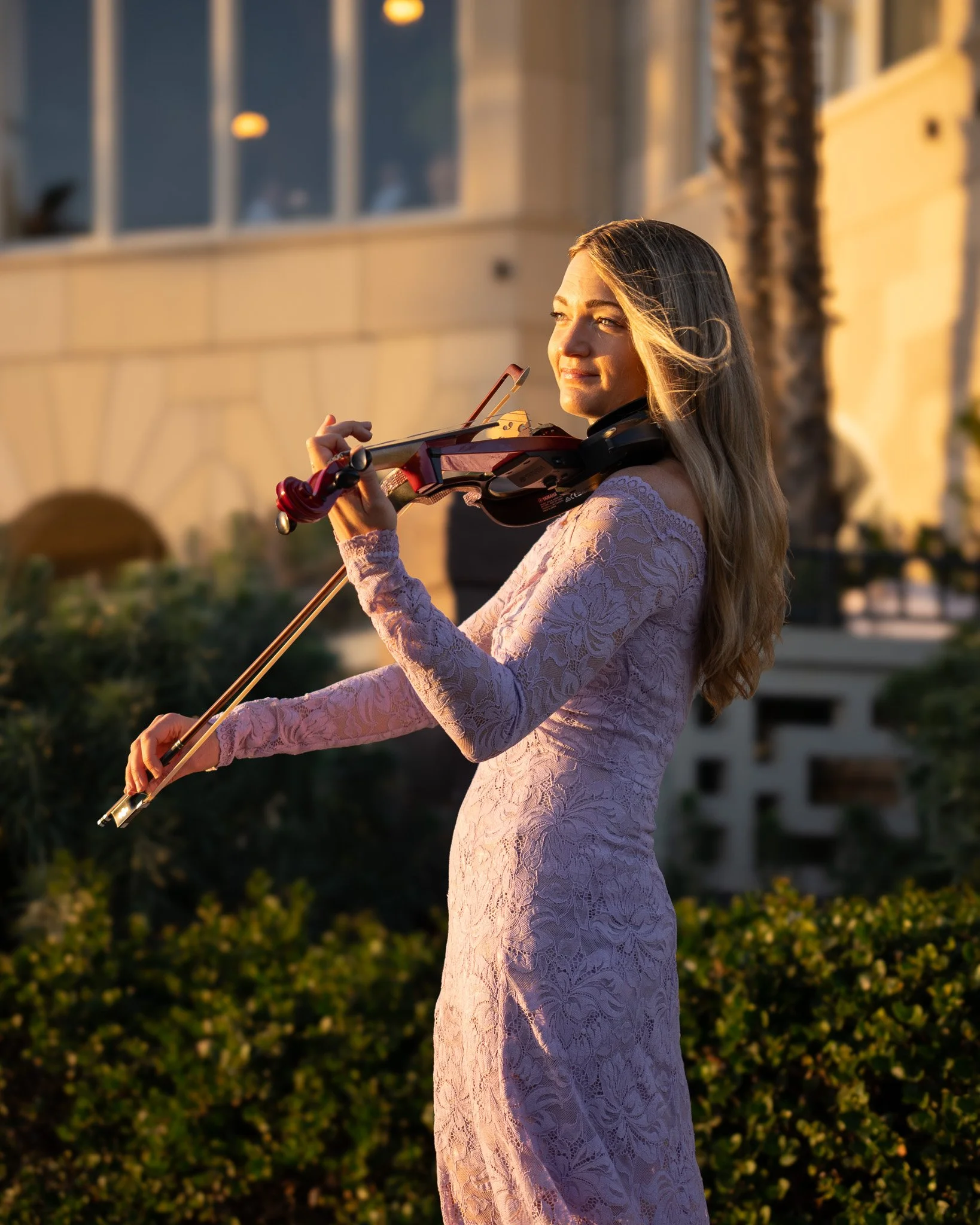 A woman in a pink lace dress playing the violin outdoors during sunset.