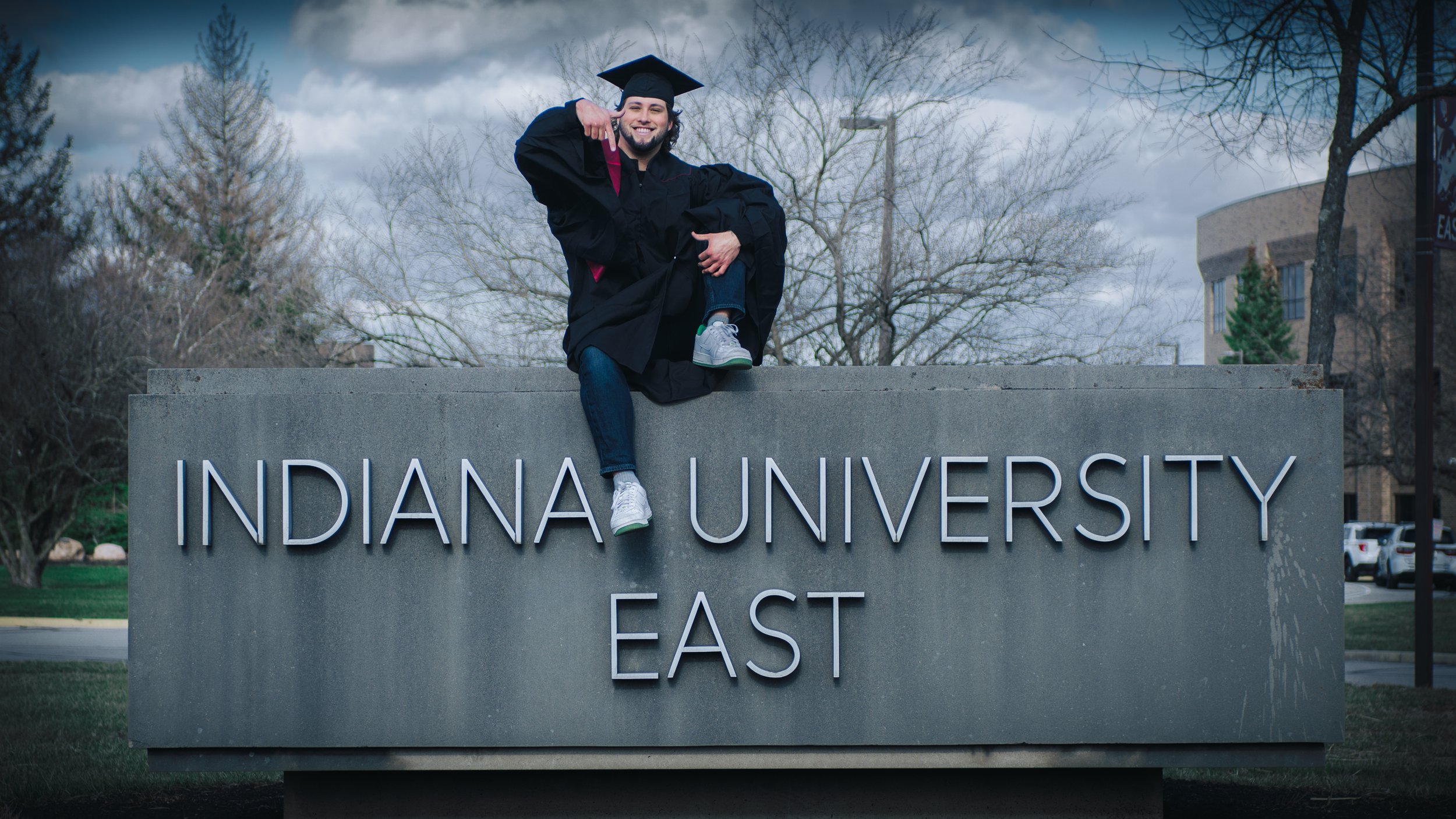 A graduate in cap and gown sitting on a sign that reads 'Indiana University East' with trees and buildings in the background