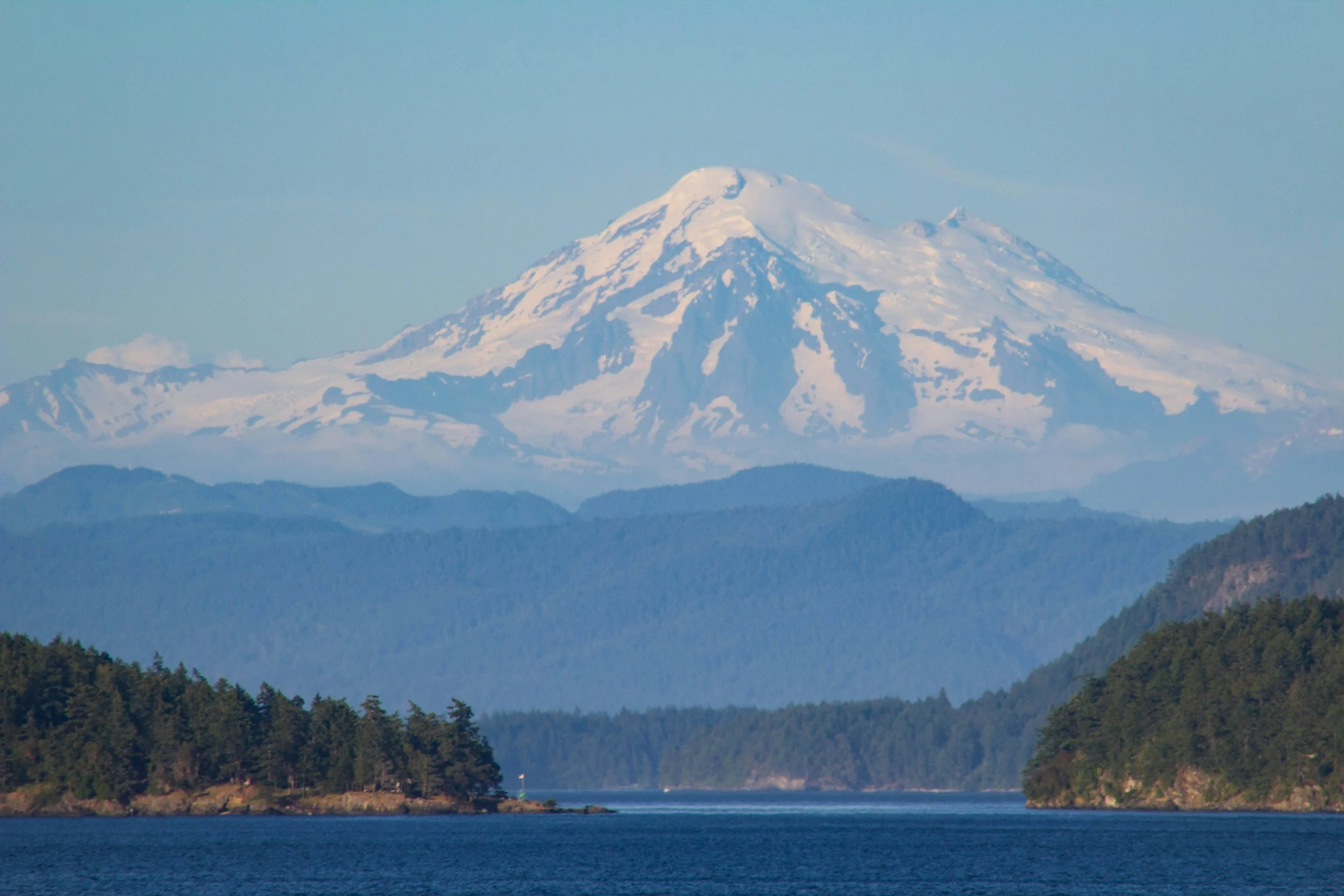Snow-capped mountain peak rising above a forested landscape and a body of water in the foreground.